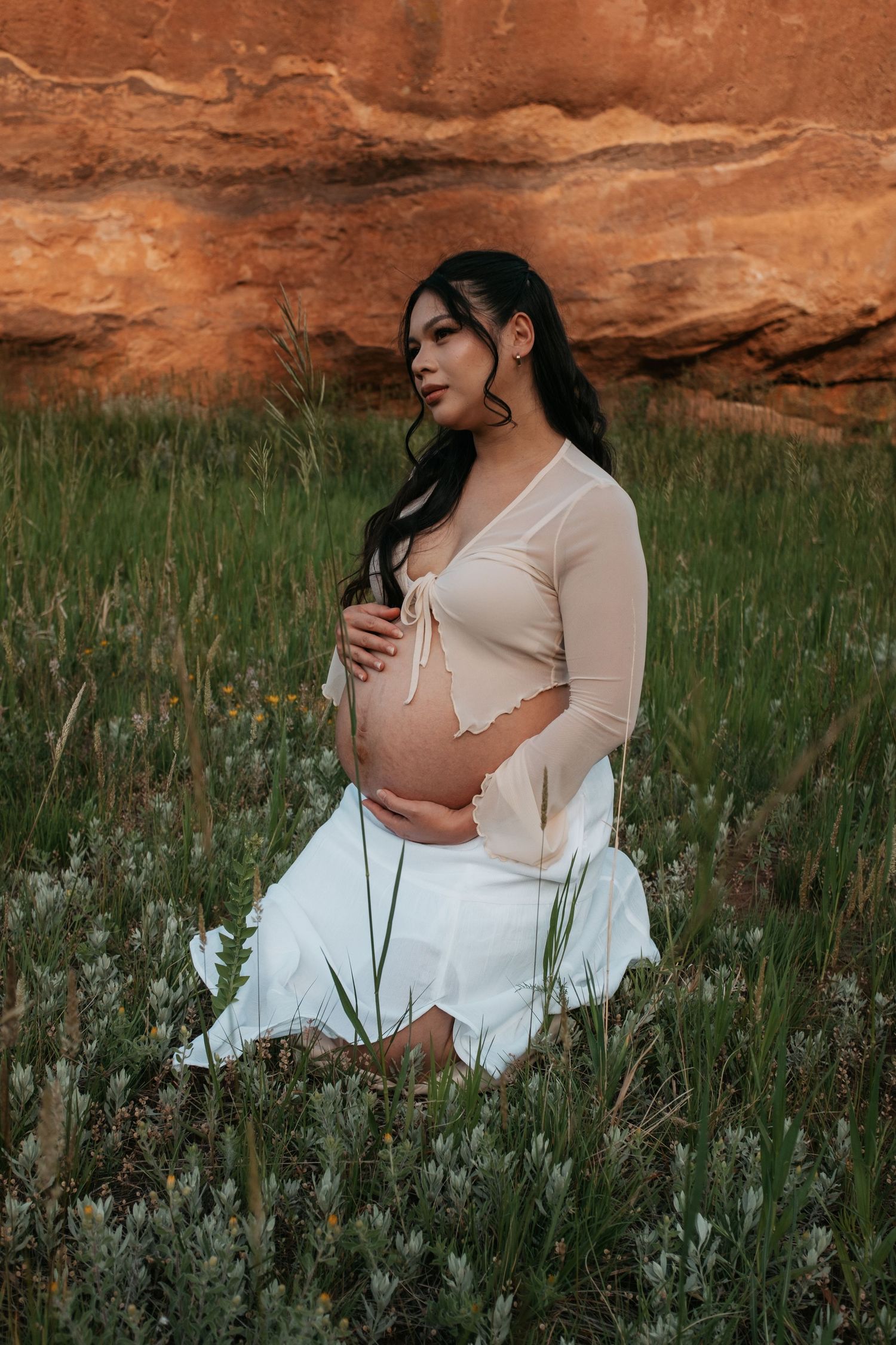 A maternity photo of someone in a white dress sitting in a wildflower field against red rocks at sunset.