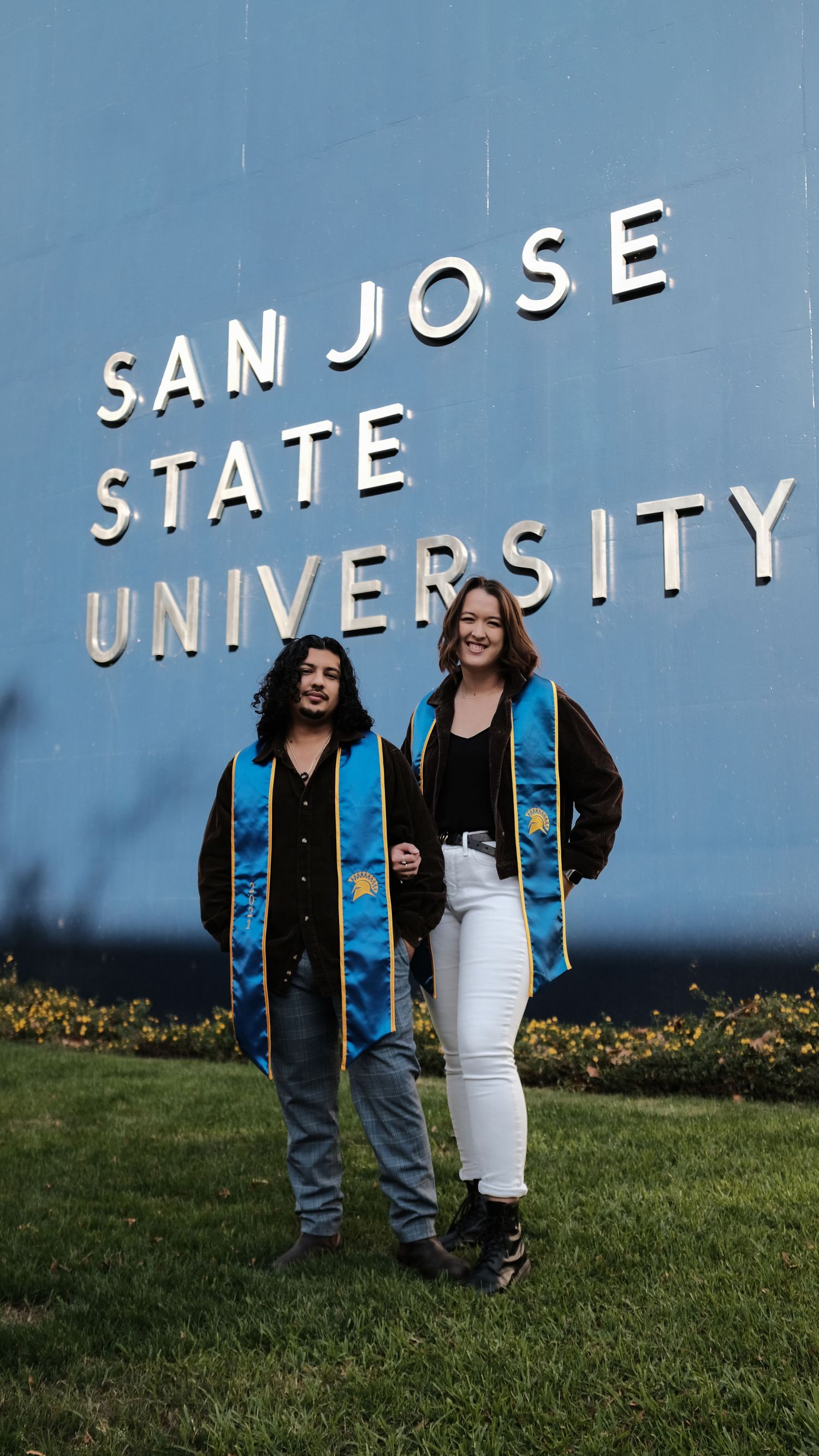 Two graduates in graduation stoles stand in front of San Jose State University sign.