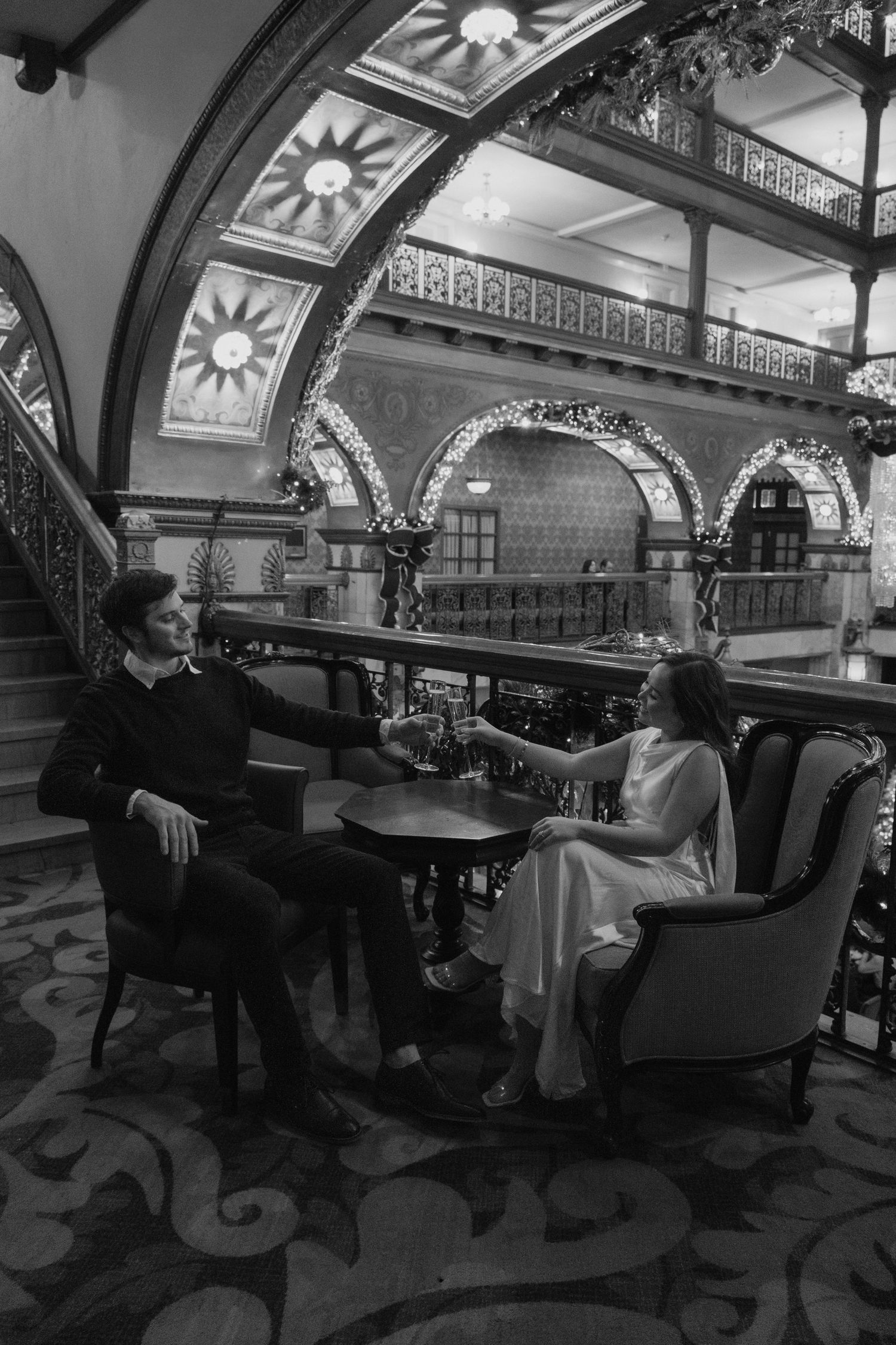 Couple sitting in elegant chairs in ornate historic hotel lobby, black and white photo.