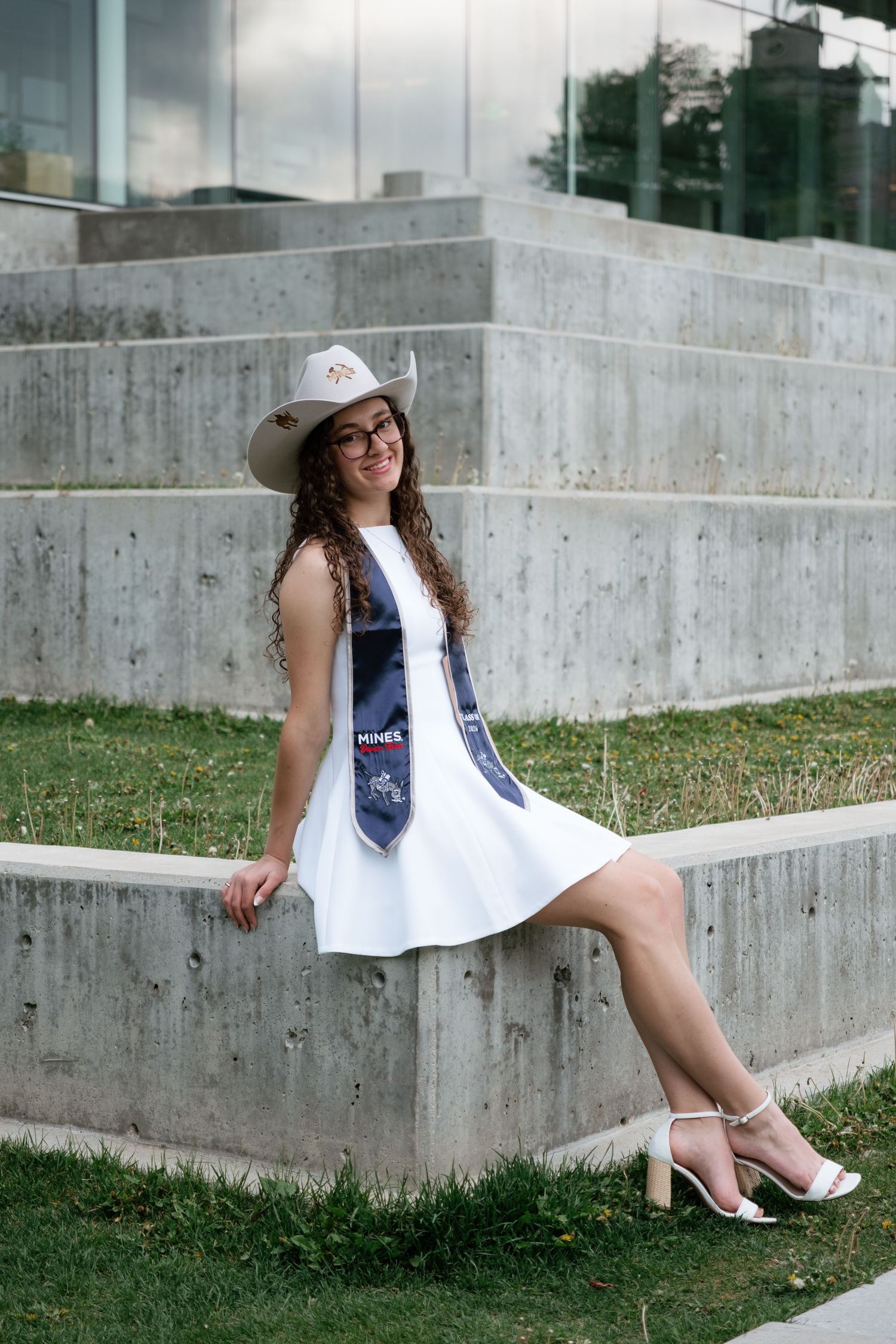 Young woman in white dress and cowboy hat wearing a graduation stole sits on concrete steps outdoors.