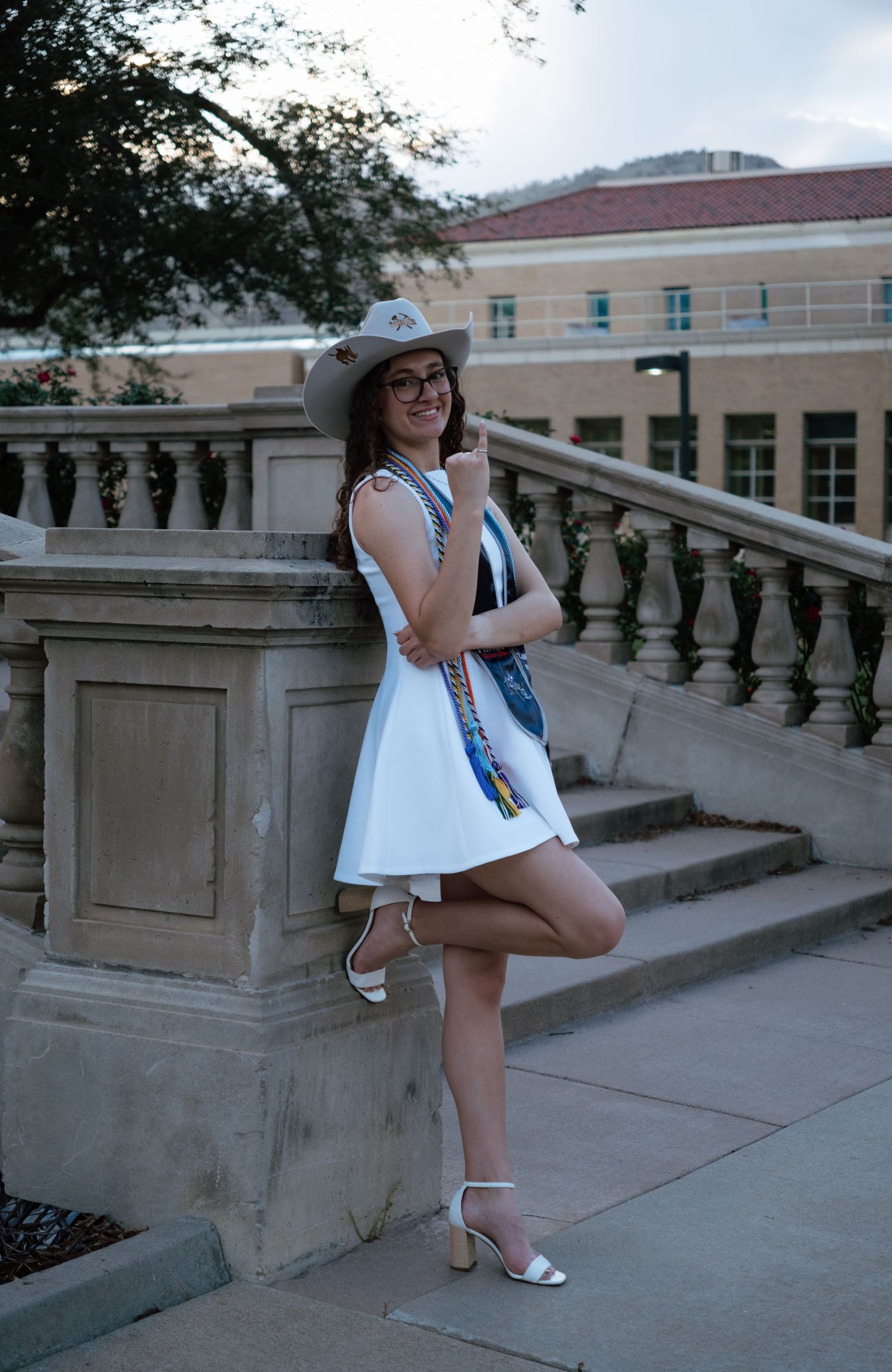 Young woman in white dress, wide-brim hat, and heels poses by stone balustrade on sunny campus steps.