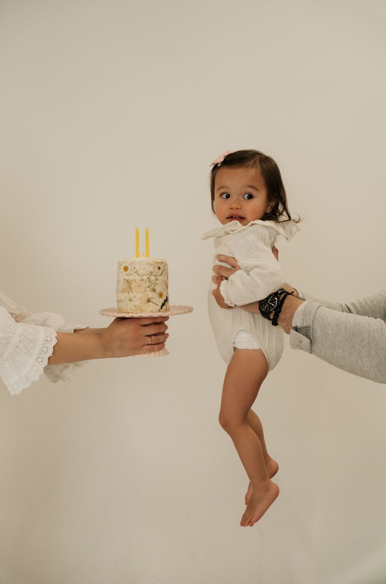 A baby in a white outfit is held up by an adult near a small birthday cake with candles on a light background.