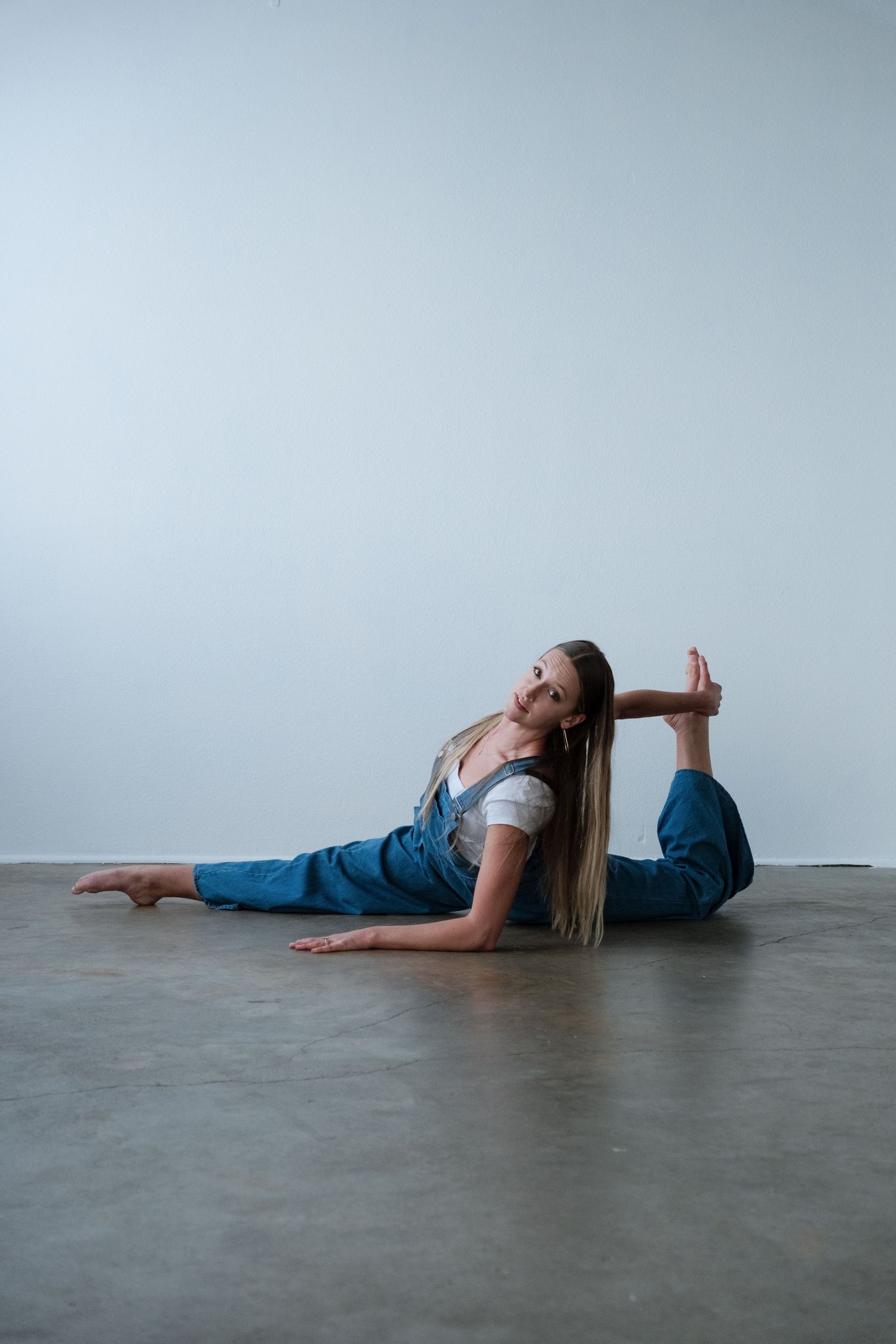 Flexible woman in blue overalls performing a split stretch on studio floor, holding her foot while looking at camera.