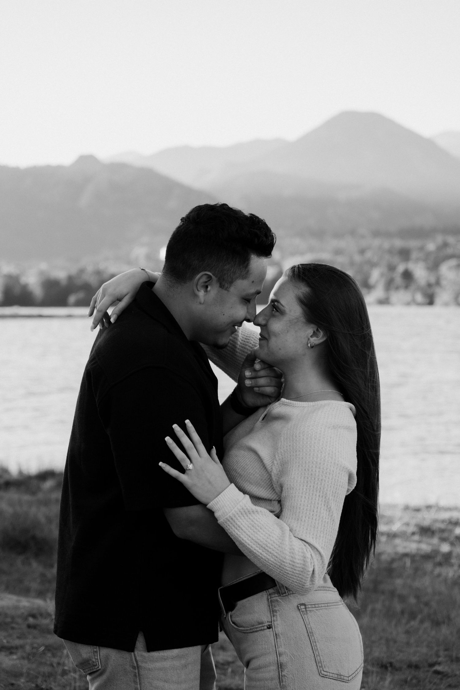 Black and white photo of a romantic couple embracing near a lake with mountains in the background.