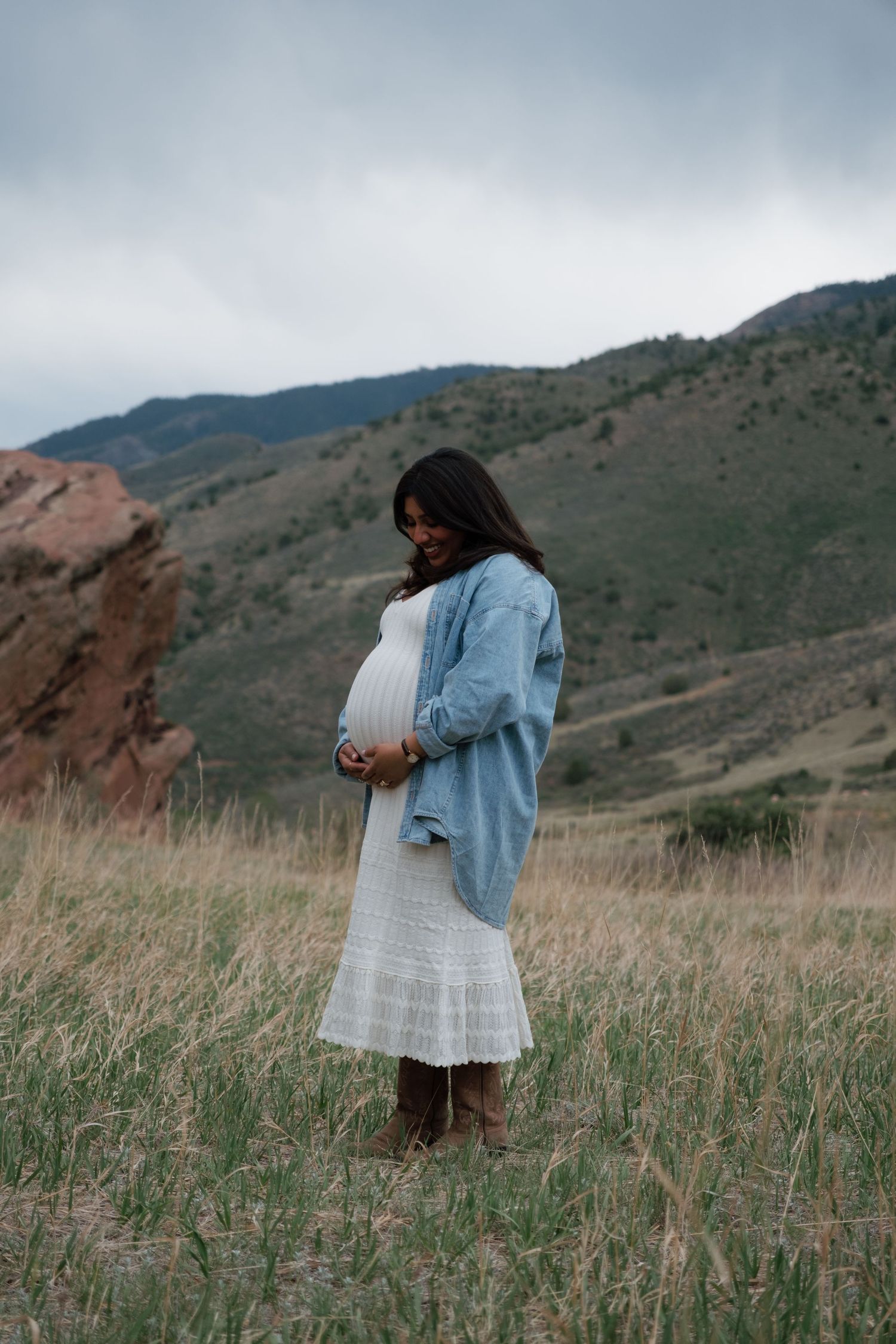 Pregnant woman in white dress and denim jacket stands in mountain meadow, cradling her baby bump on a cloudy day.