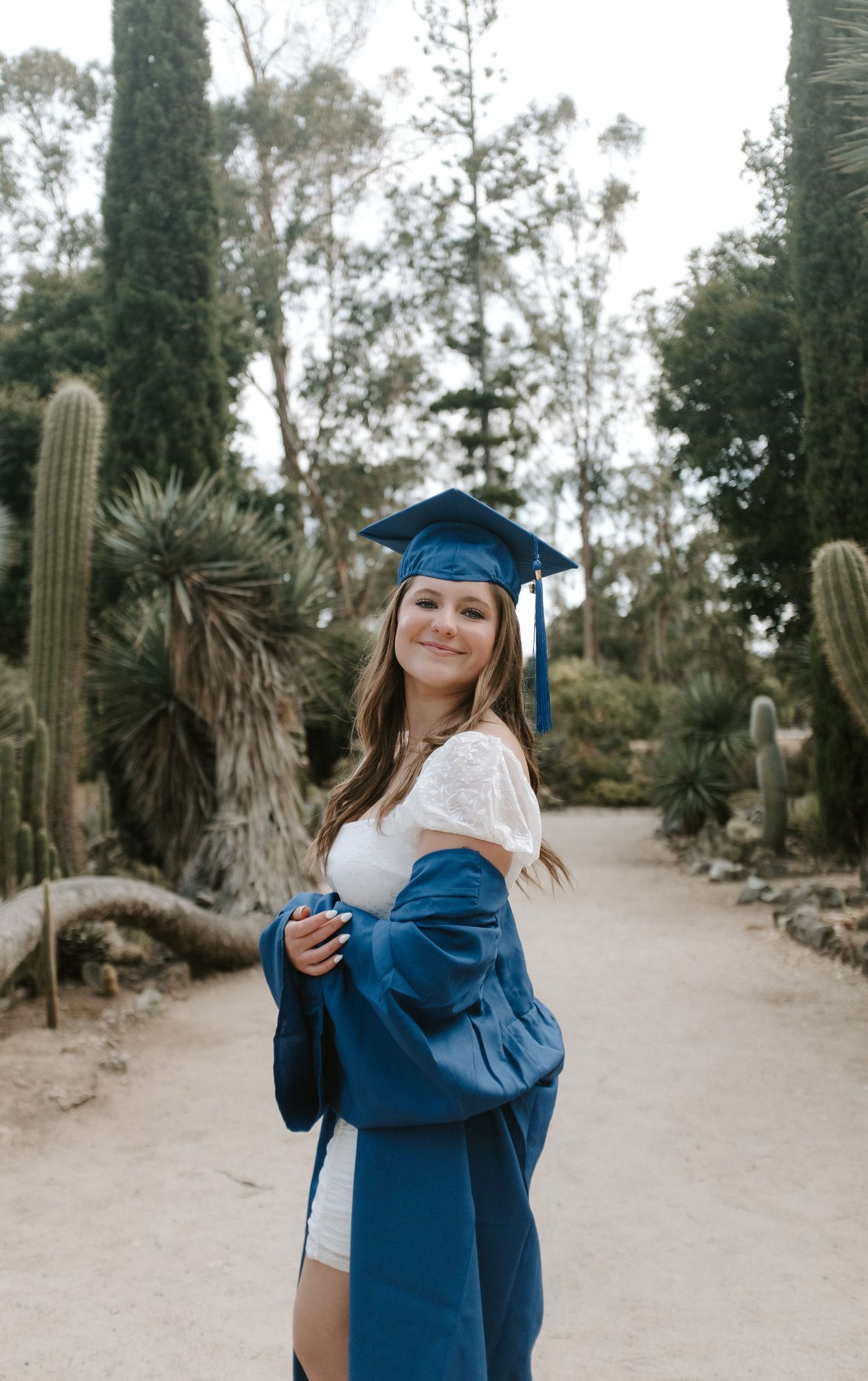 Graduate in blue cap and gown poses joyfully in an outdoor garden setting with palm trees and cacti.