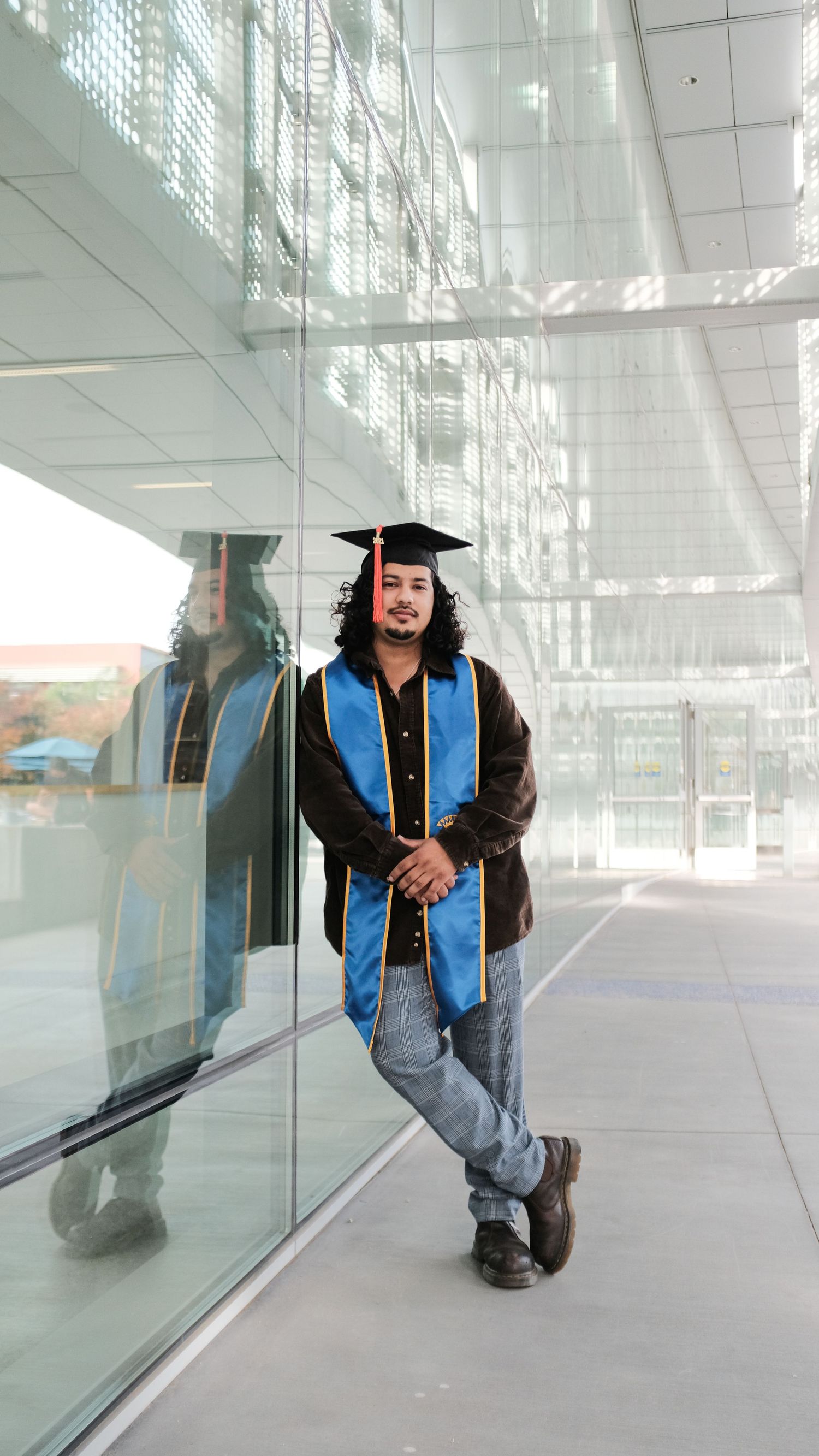 Graduate in blue stole and black gown poses artistically against reflective glass building.