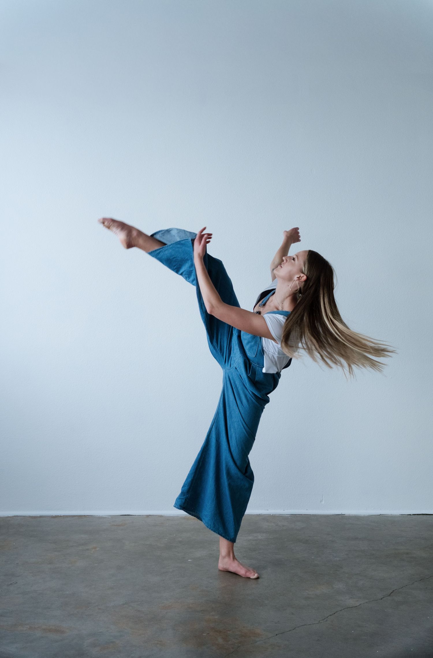A barefoot dancer in a blue jumpsuit performs a high kick while balancing gracefully in a studio.