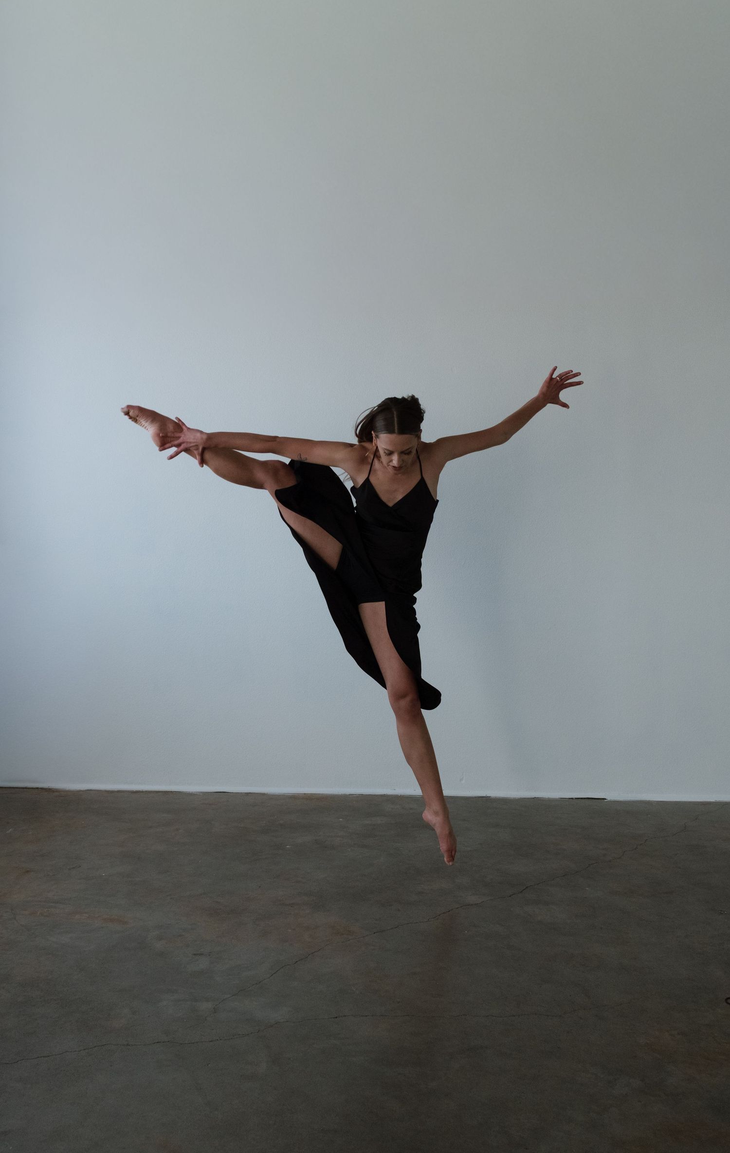 A dancer in black dress leaps gracefully mid-air, one leg extended high, arms outstretched in a modern dance pose.