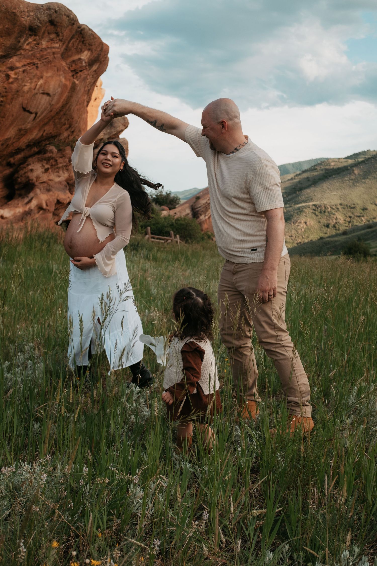 Pregnant woman in white dress dances with bald man outdoors as young child watches near red rocks.