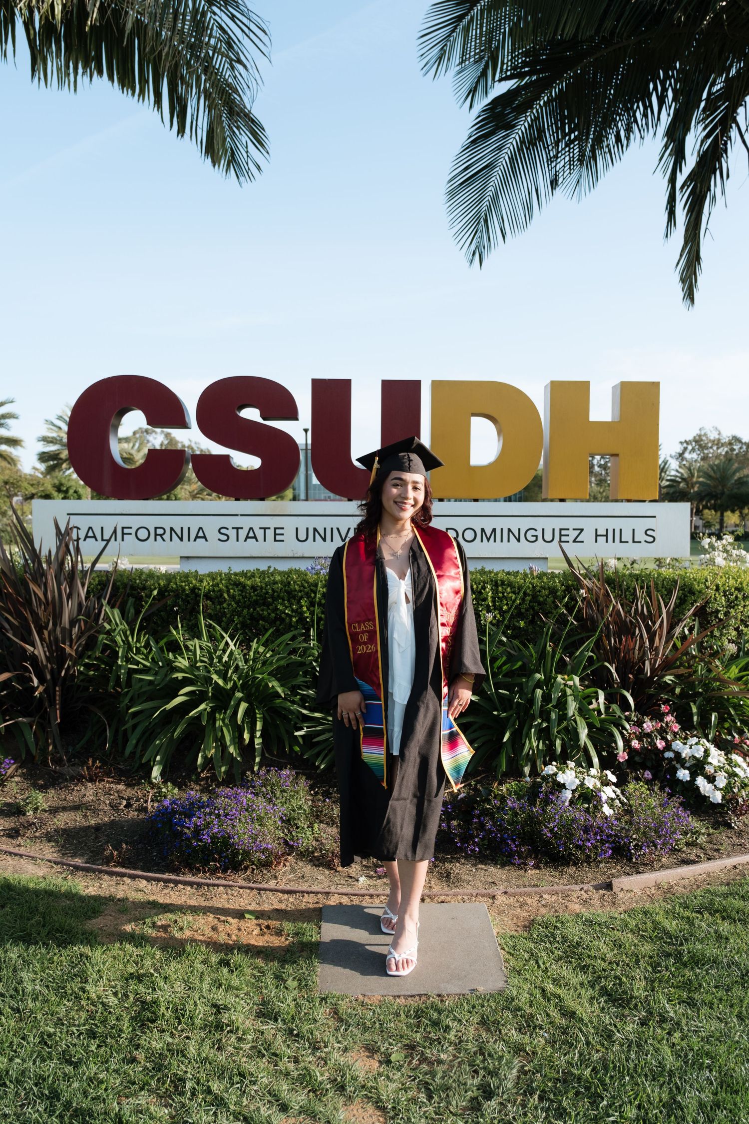 A smiling graduate in cap and gown poses in front of the CSUDH California State University Dominguez Hills sign.