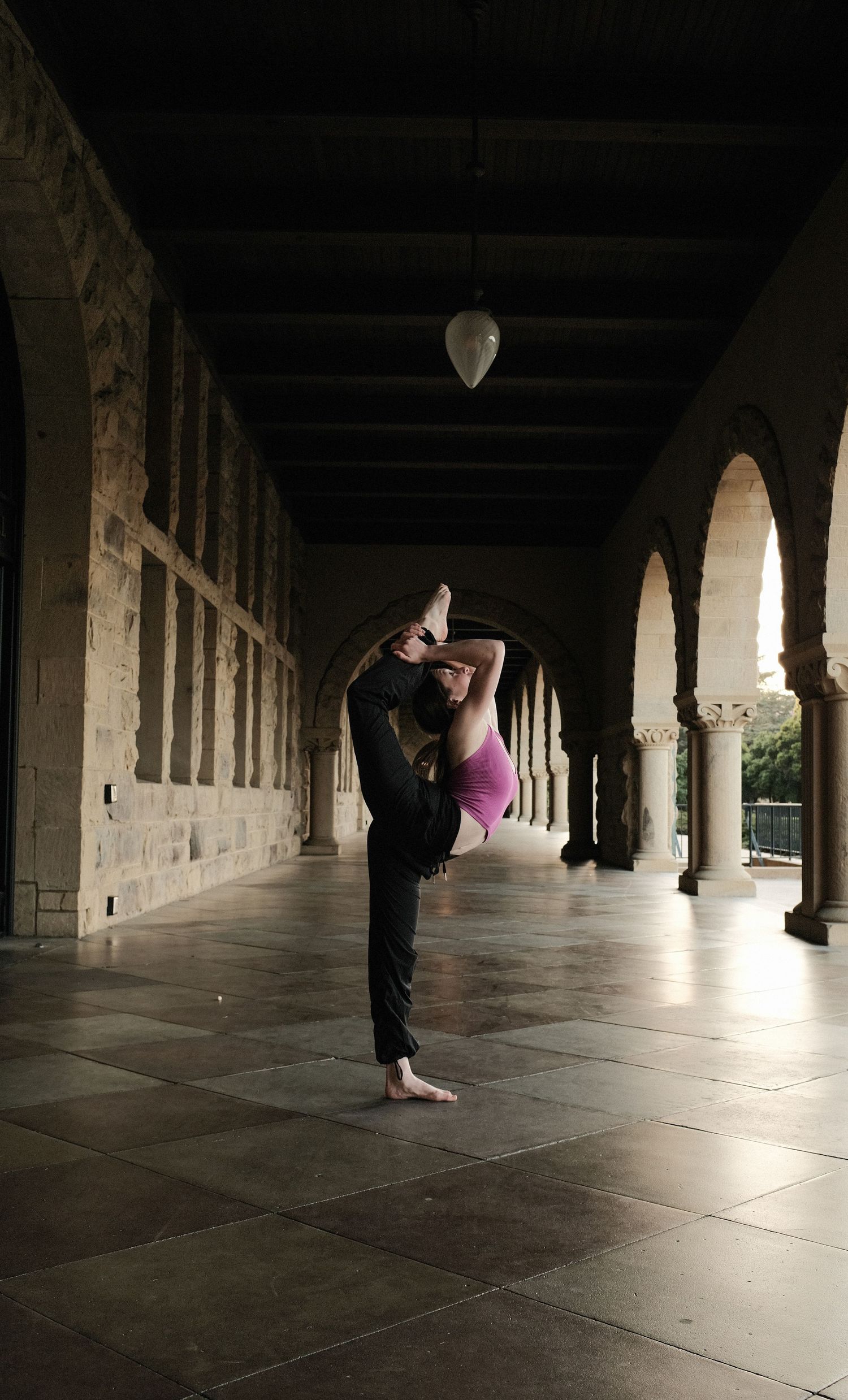 A dancer performs a graceful pose in a historic colonnade with arched pillars and dramatic lighting.