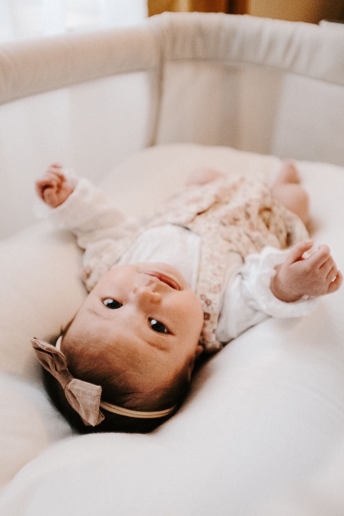 A baby in lace clothing lies on their back in a white crib.