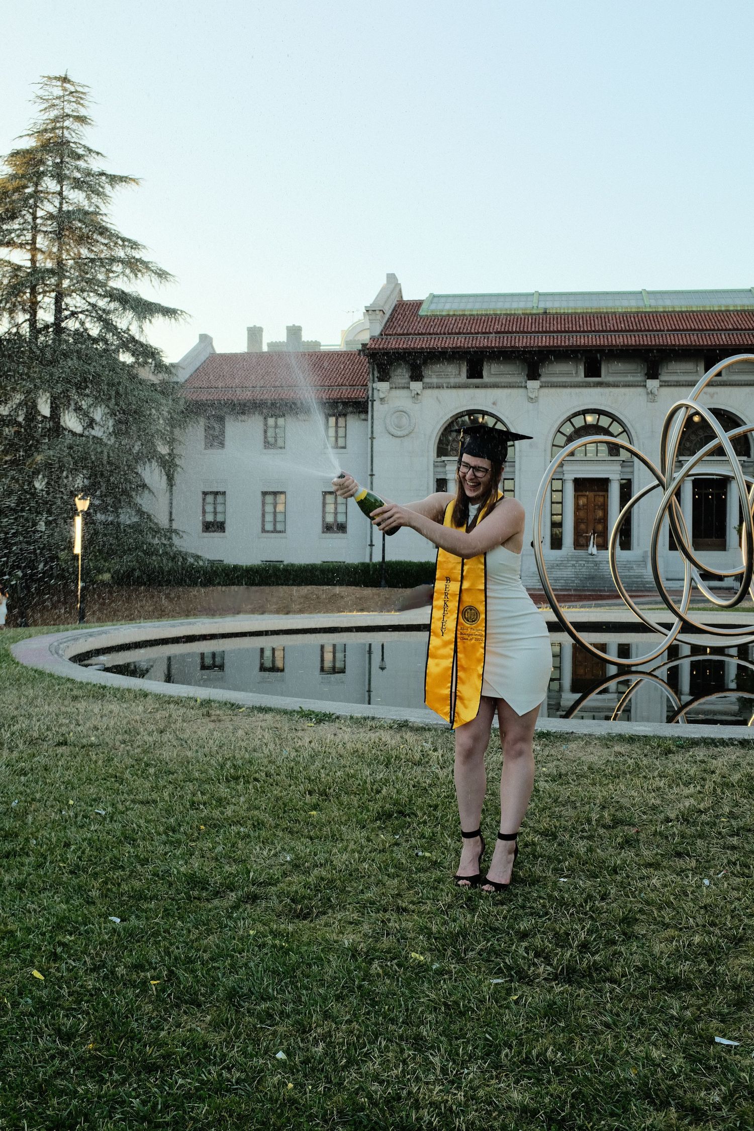Graduate wearing white dress and yellow sash poses with water fountain and historic building in background.