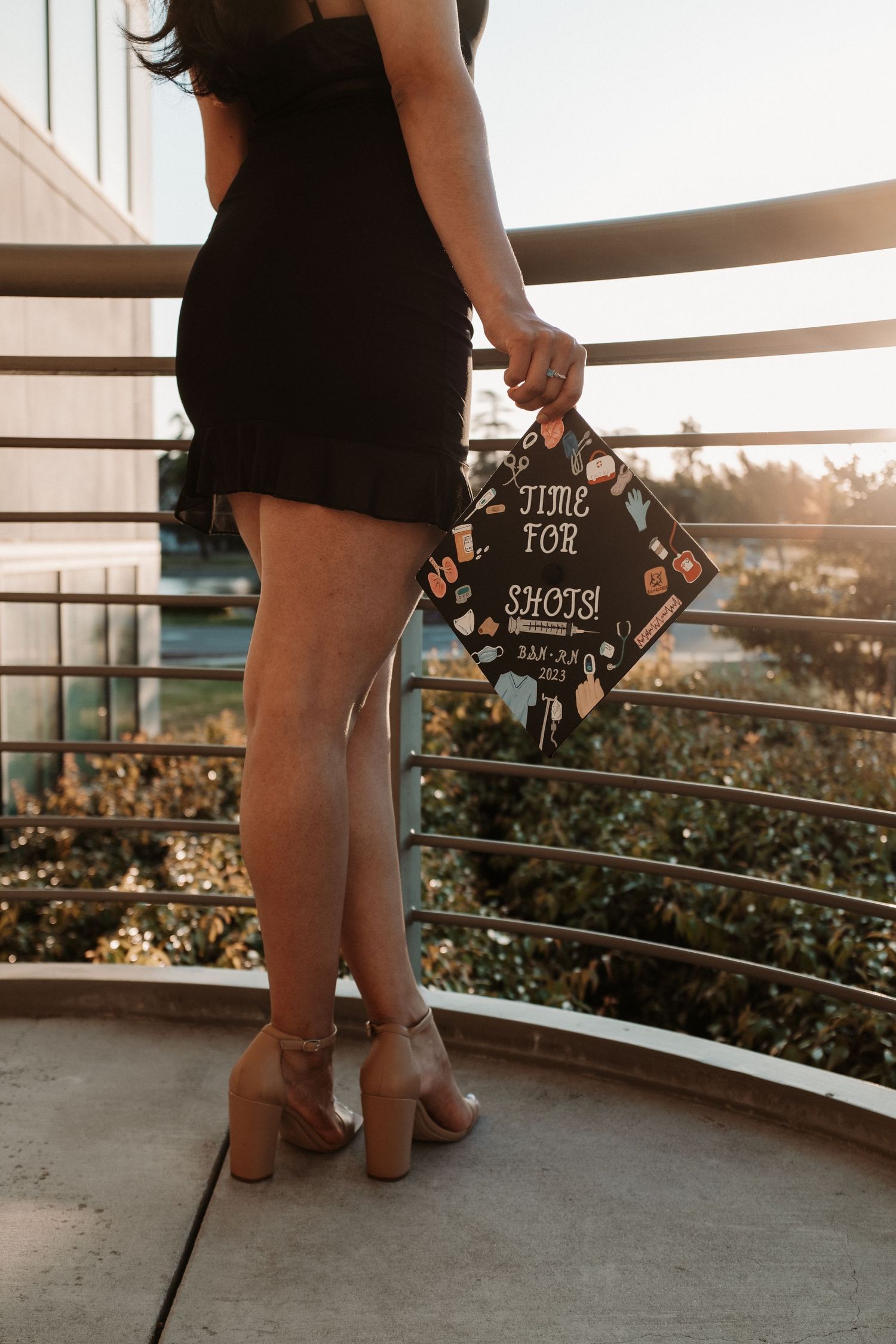 Graduate in black dress holding decorated cap against sunset on balcony.