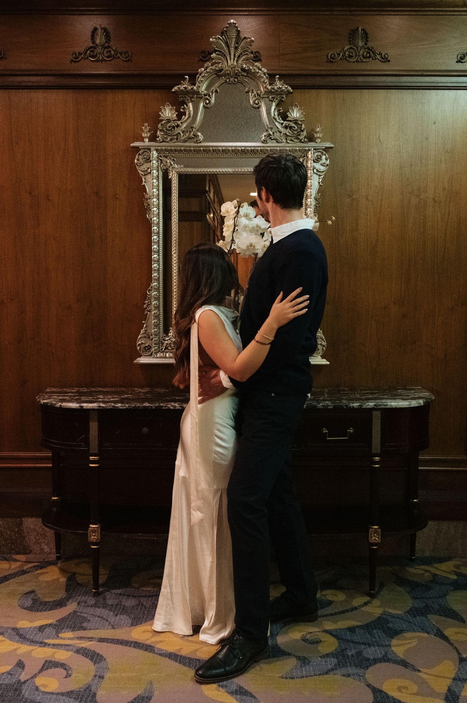 Couple embracing in front of ornate vintage mirror on wooden console table in elegant hotel lobby.