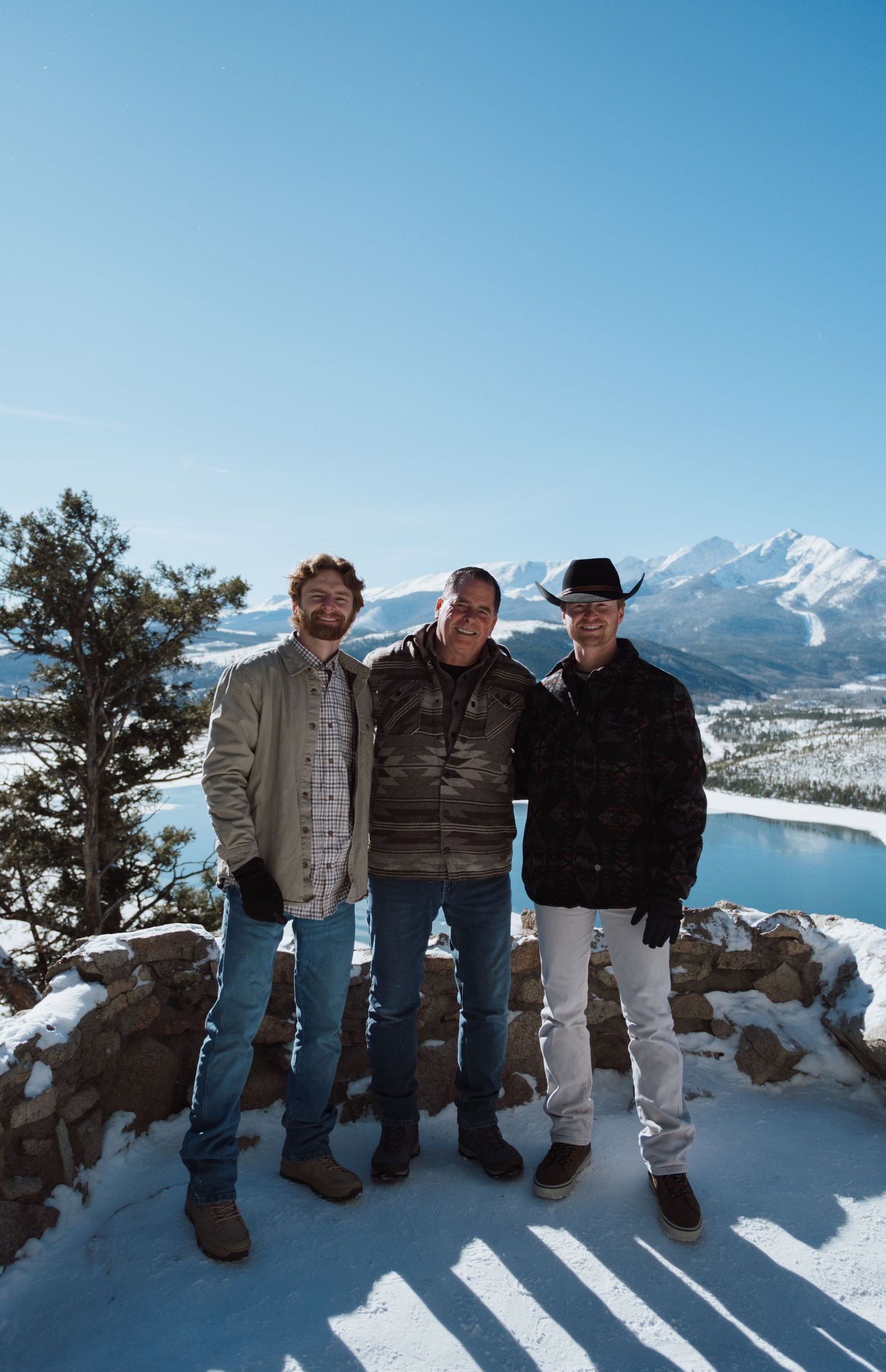 Three men pose smiling at a snowy mountain overlook with a frozen lake and peaks under a clear blue sky.