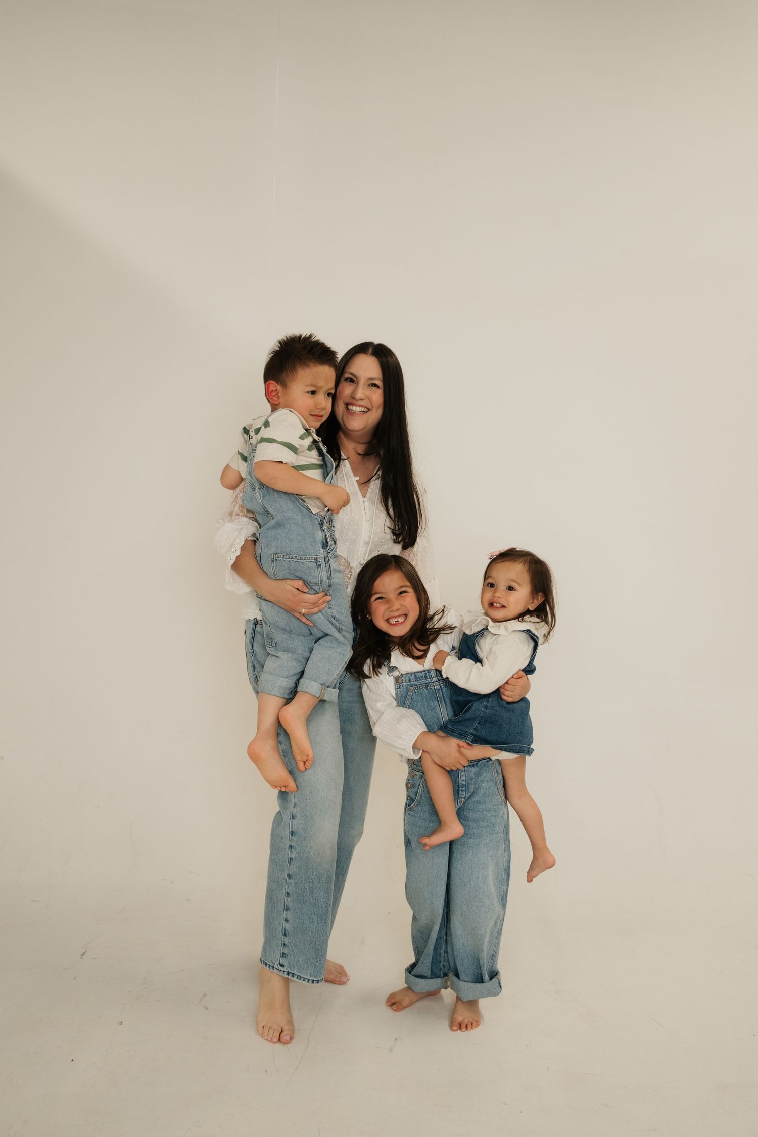 A smiling mother in jeans holds two toddlers while an older child stands beside her against a white background.