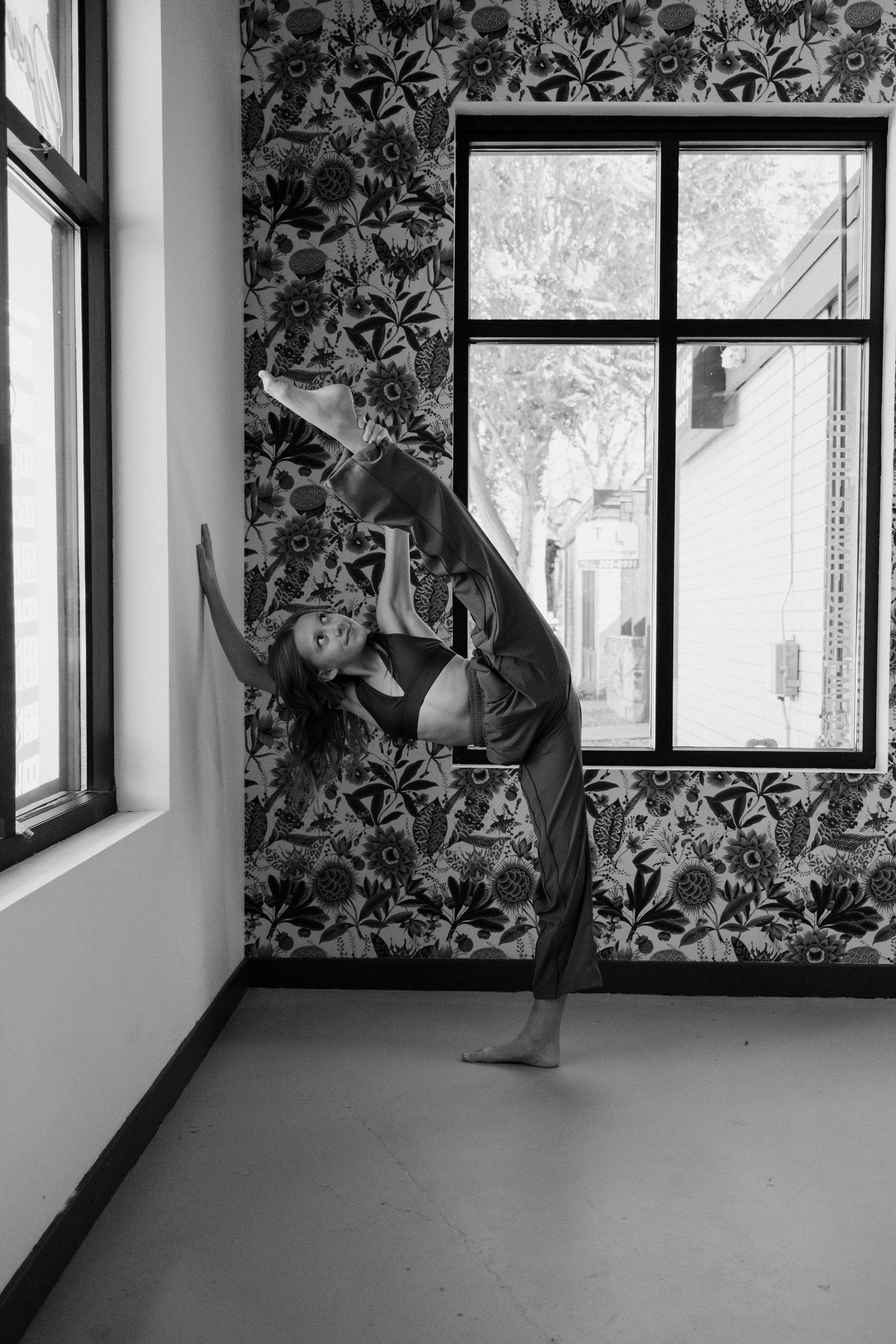 A dancer performs a graceful backbend pose in a room with floral wallpaper and large windows.