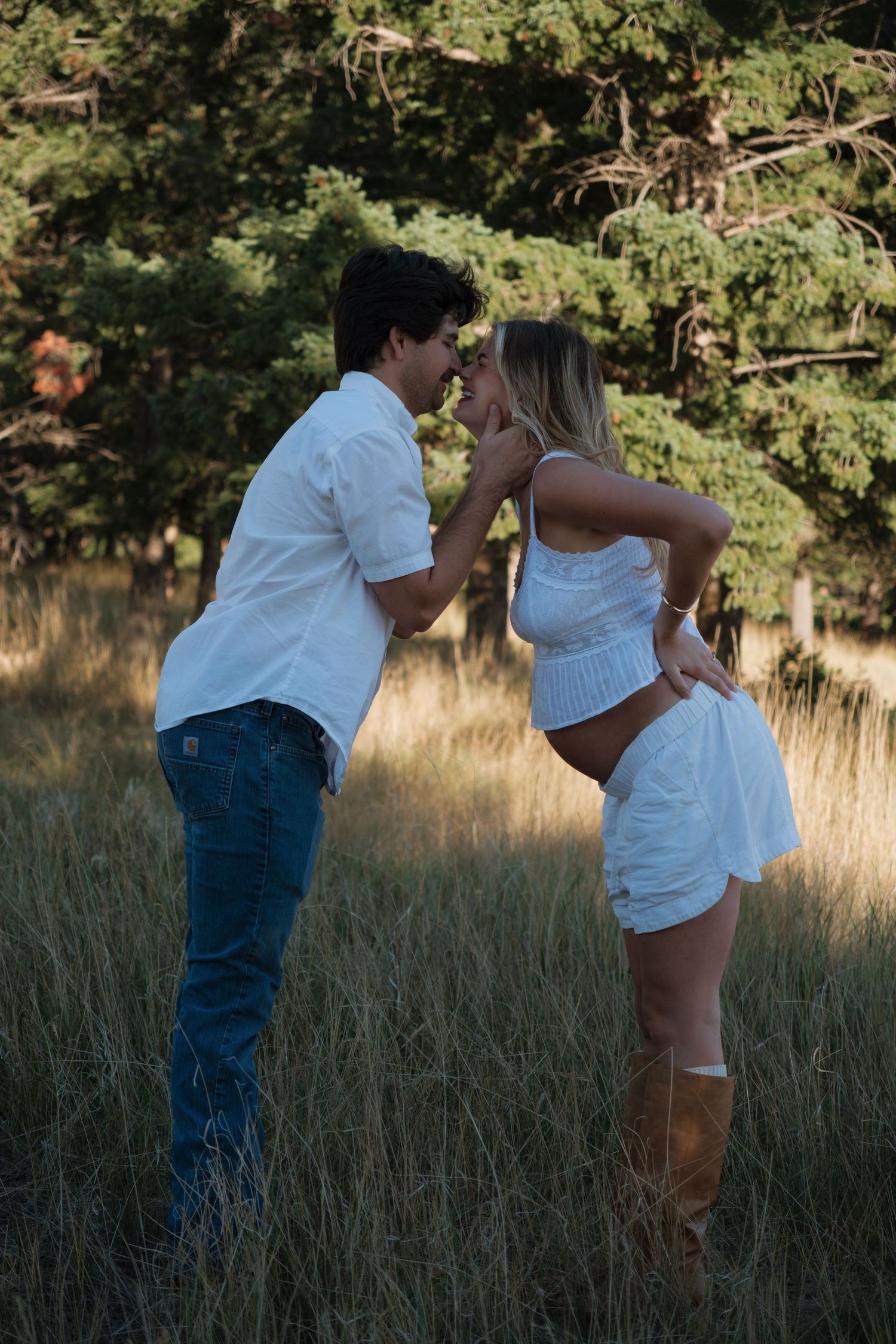 Couple in white outfits and brown boots leaning in for a kiss in a tall grass field with evergreen trees at sunset.
