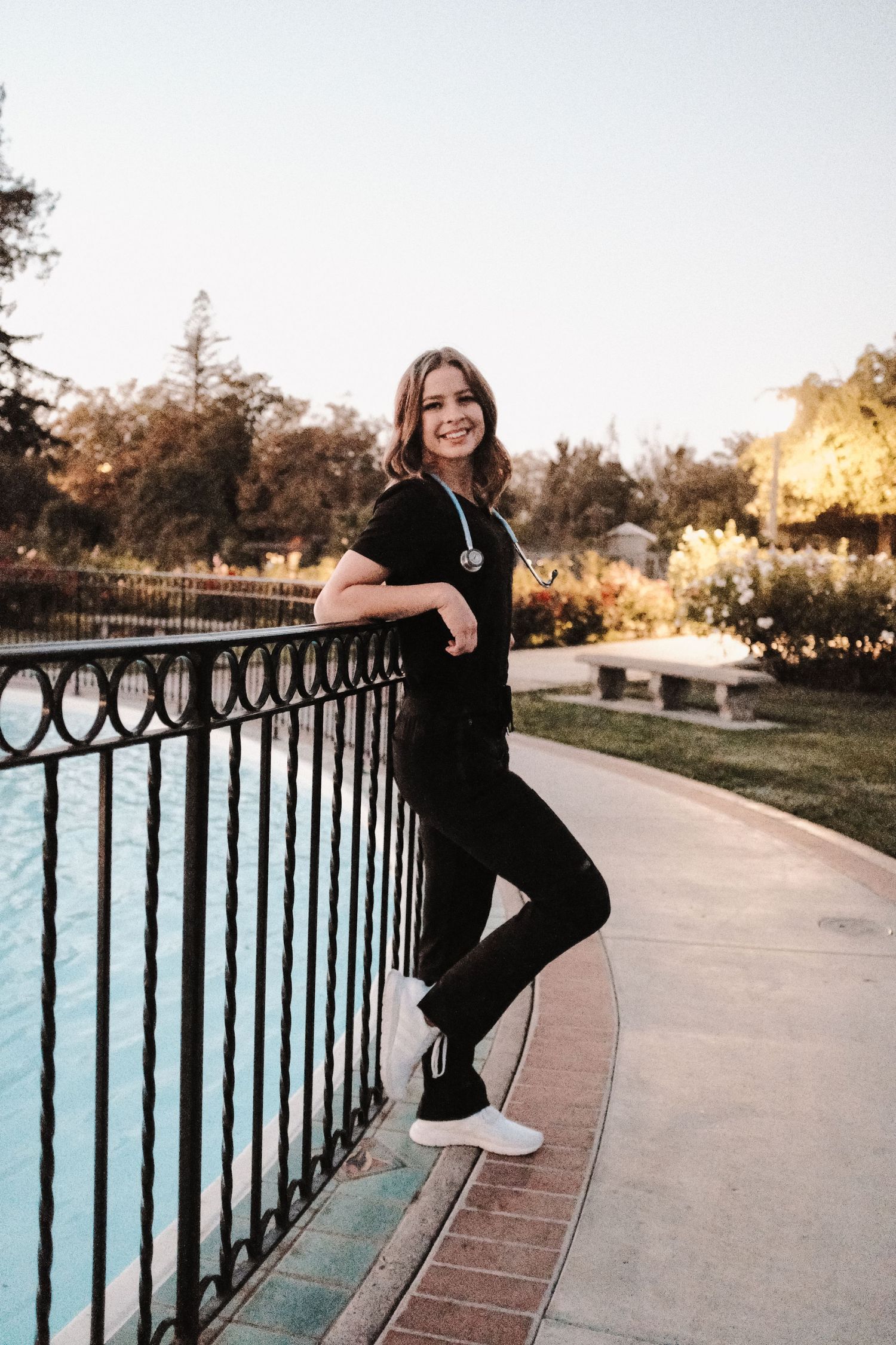 Graduate in black outfit poses against railing during golden hour.