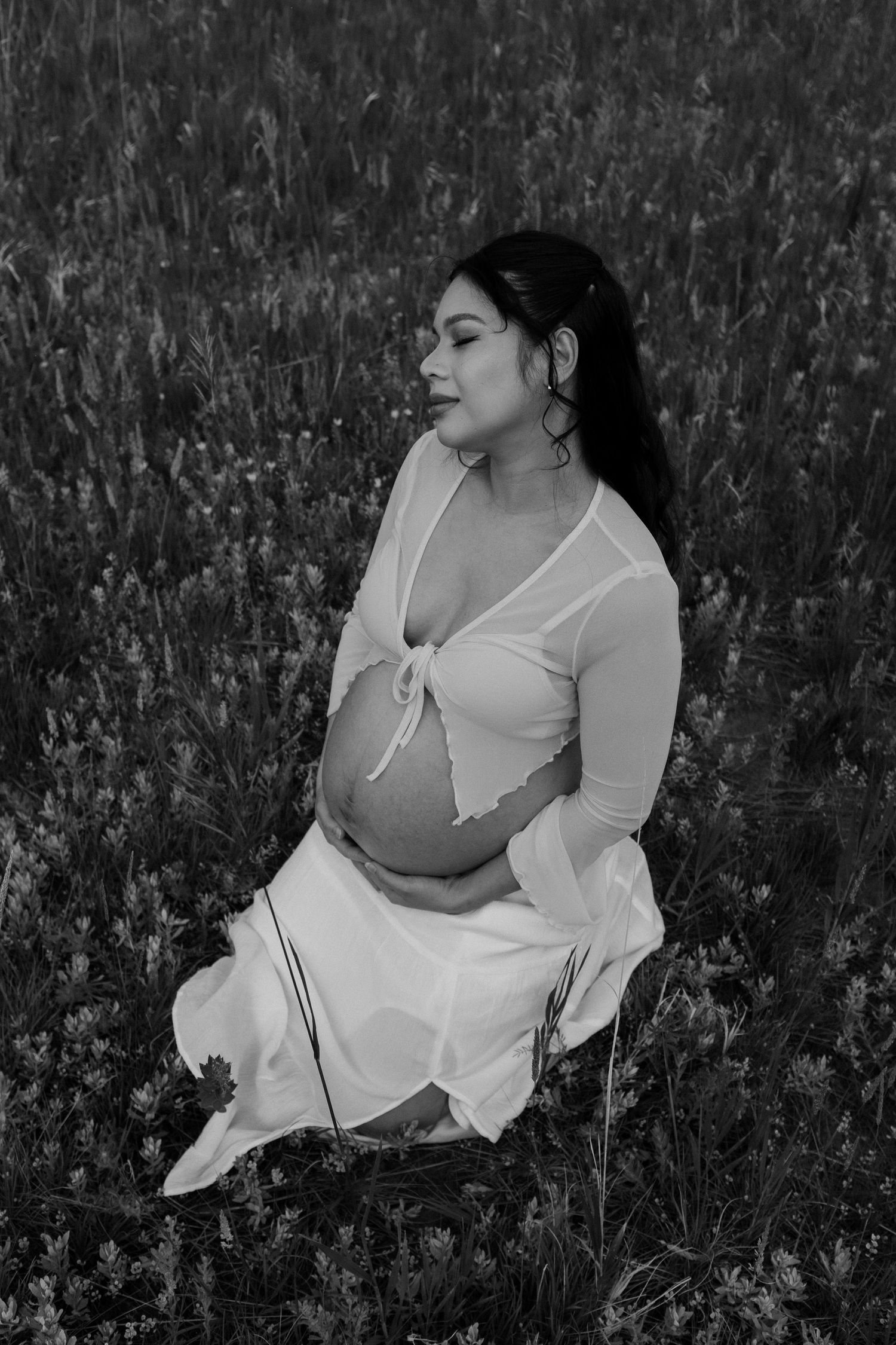 A black and white maternity portrait showing someone posed serenely among wild grasses.