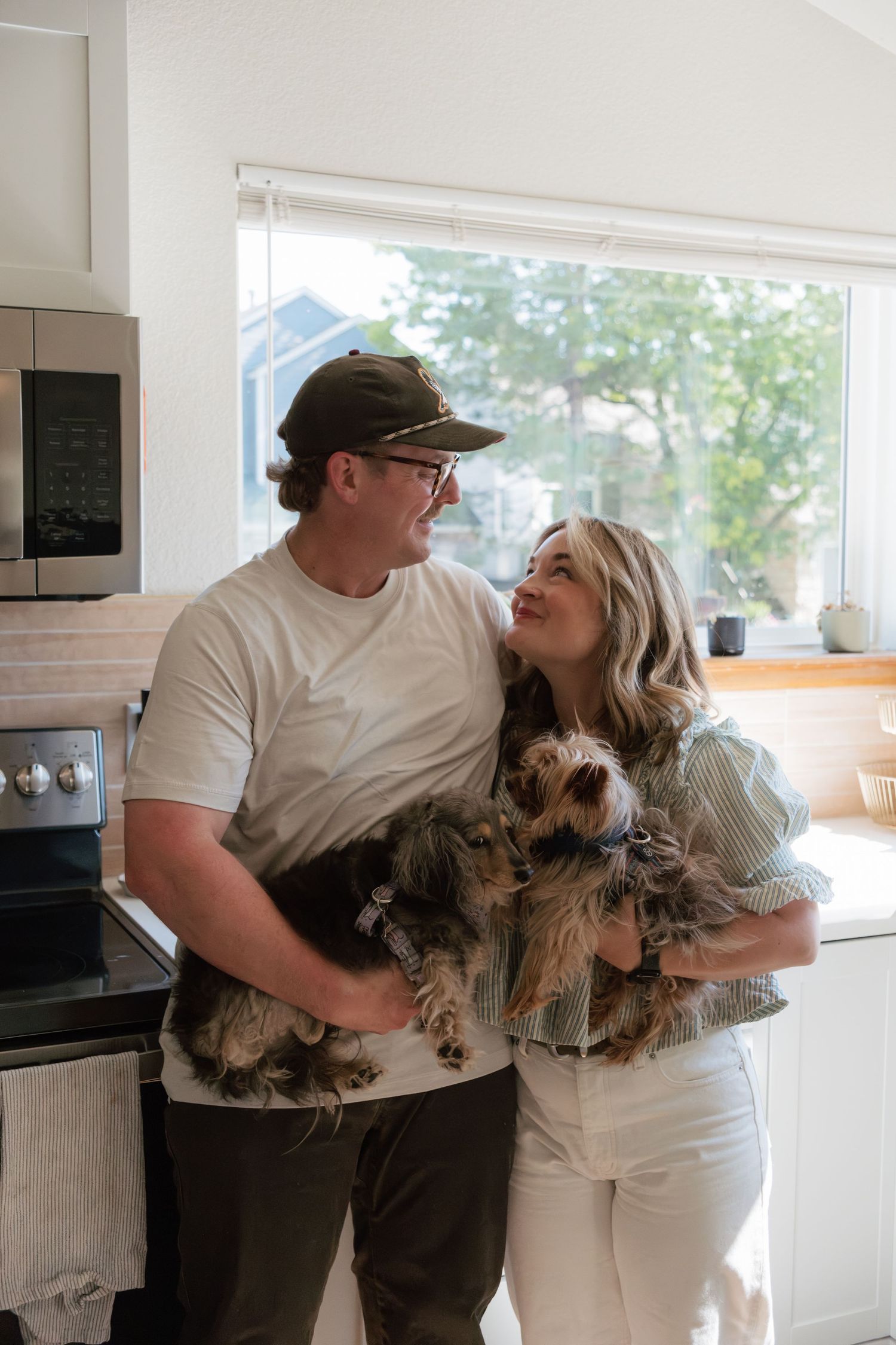 A smiling couple stands together in a bright kitchen, holding two small Yorkshire Terrier dogs while gazing happily at each other.