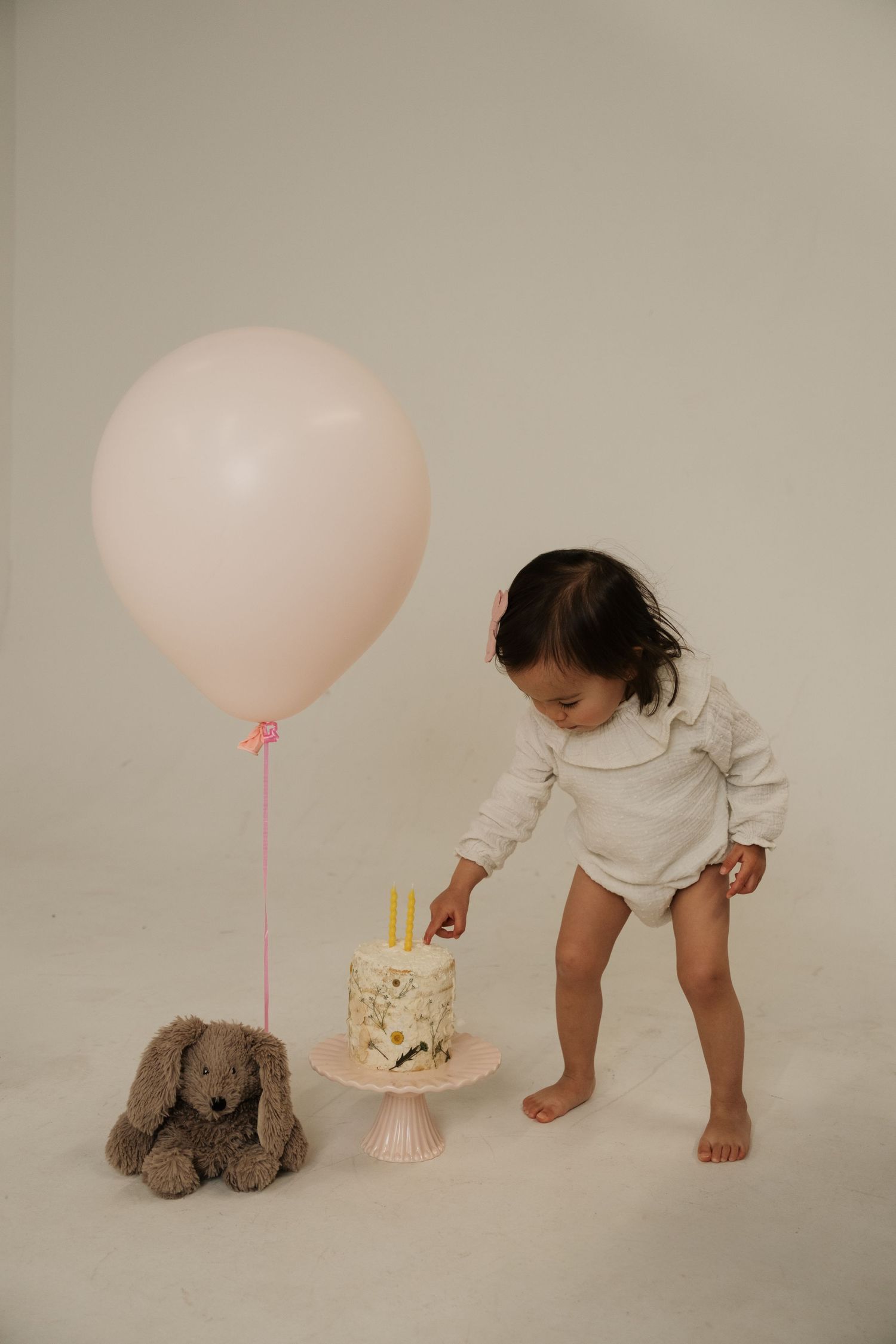 Baby girl in white onesie touching first birthday cake beside pink balloon and stuffed bunny on white background.