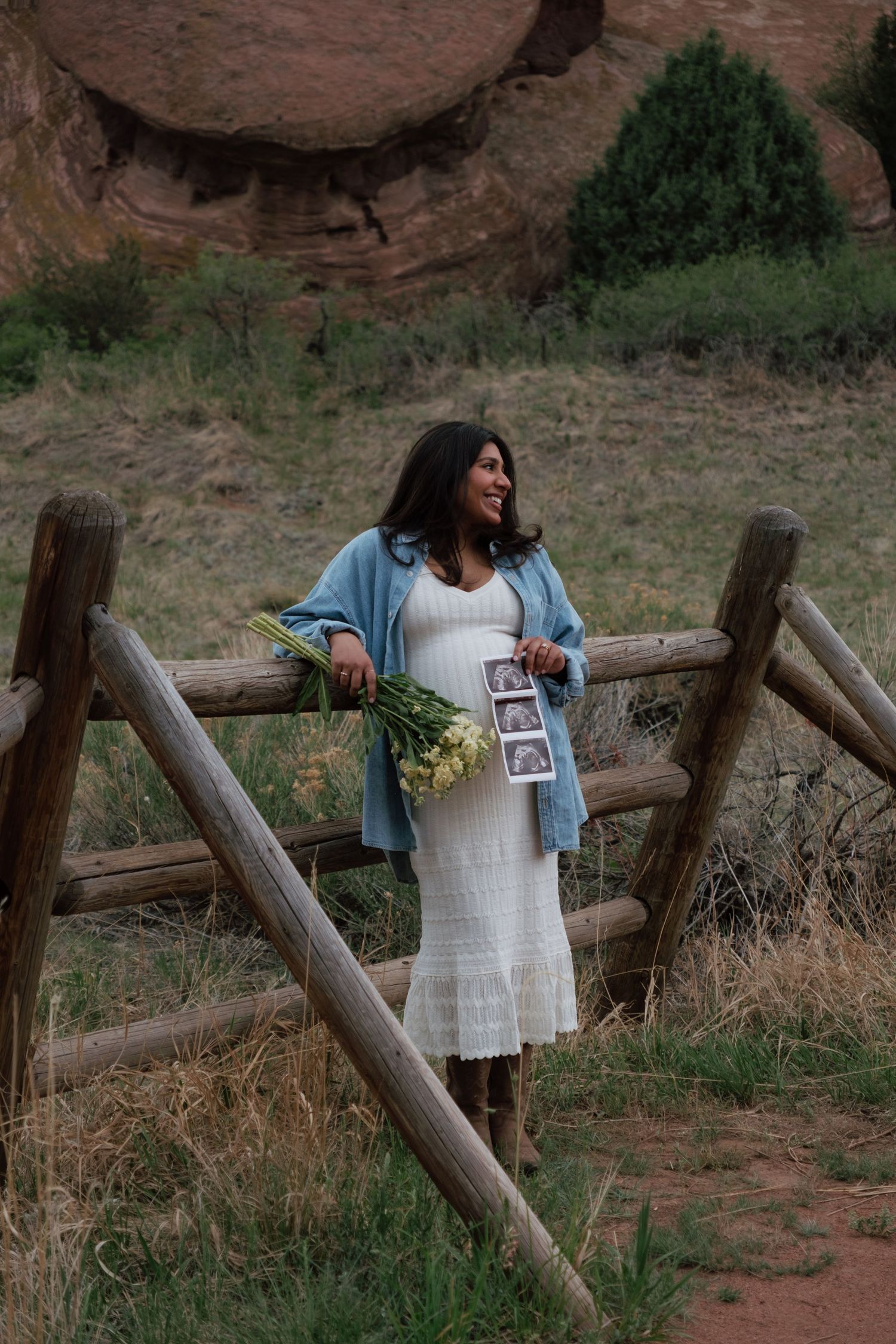 Pregnant woman in white dress and blue jacket holds flowers and ultrasound photos by a wooden fence at Red Rocks.