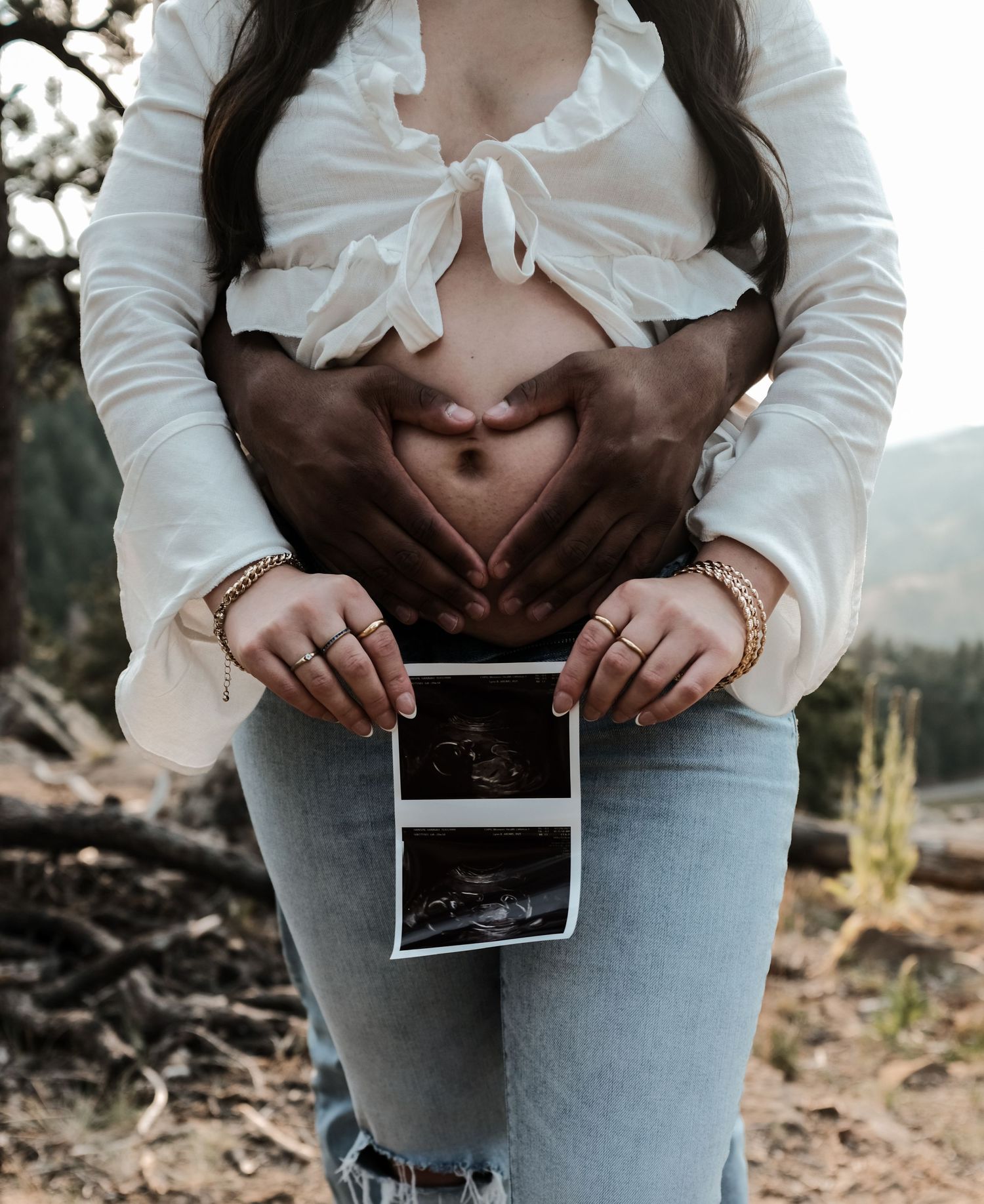 Pregnant woman holding ultrasound photo while partner forms heart shape on baby bump outdoors.