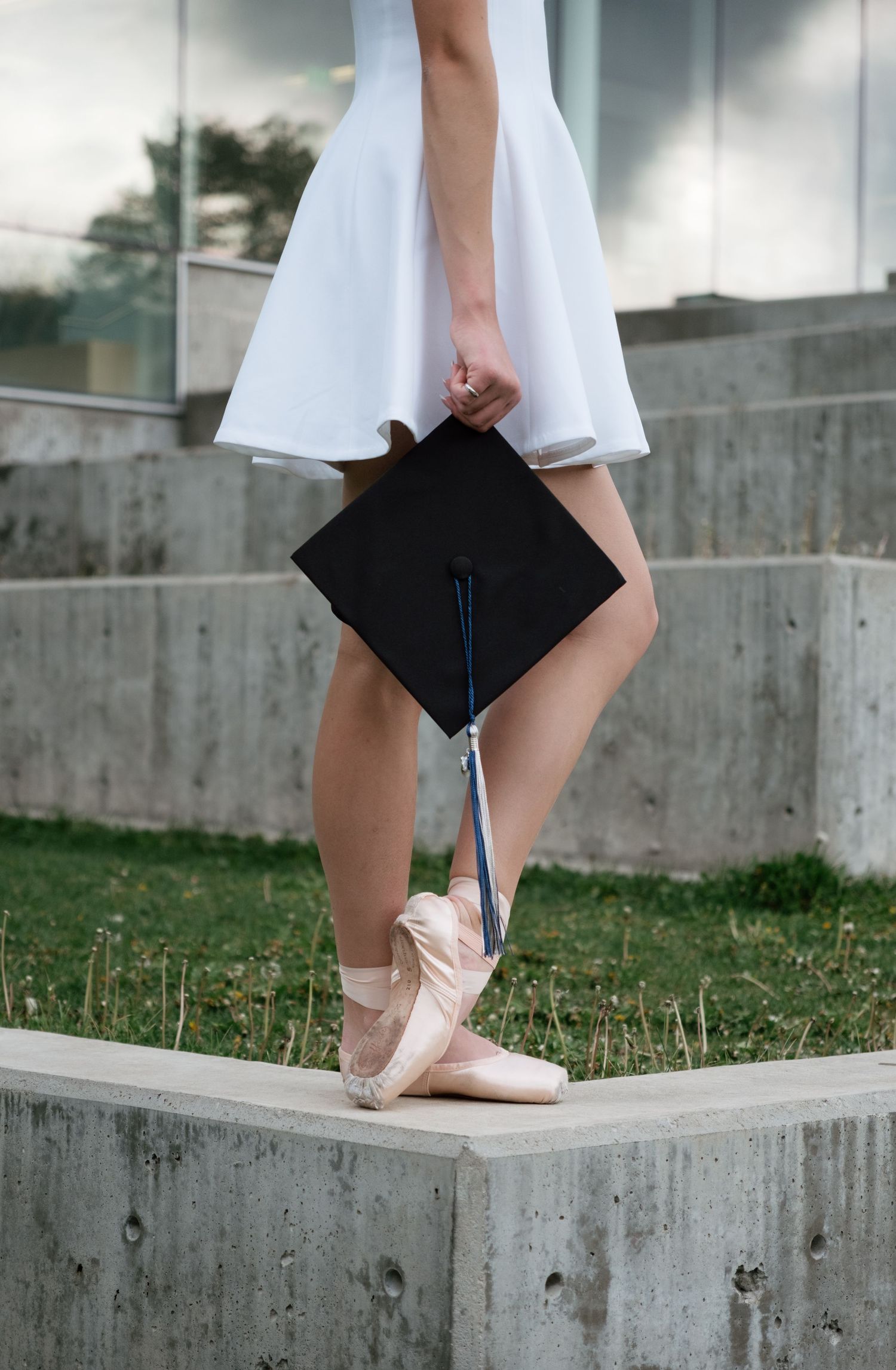 Ballerina in white dress and pointe shoes holding a black graduation cap, standing on concrete steps outdoors.