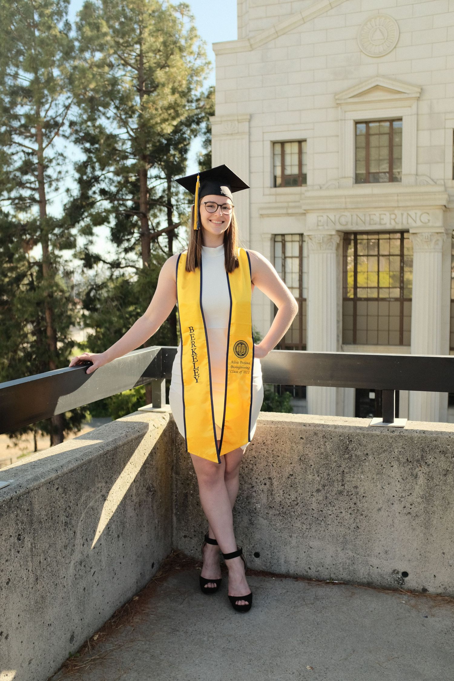 Graduate wearing yellow stole and cap poses on concrete ledge in front of academic building.