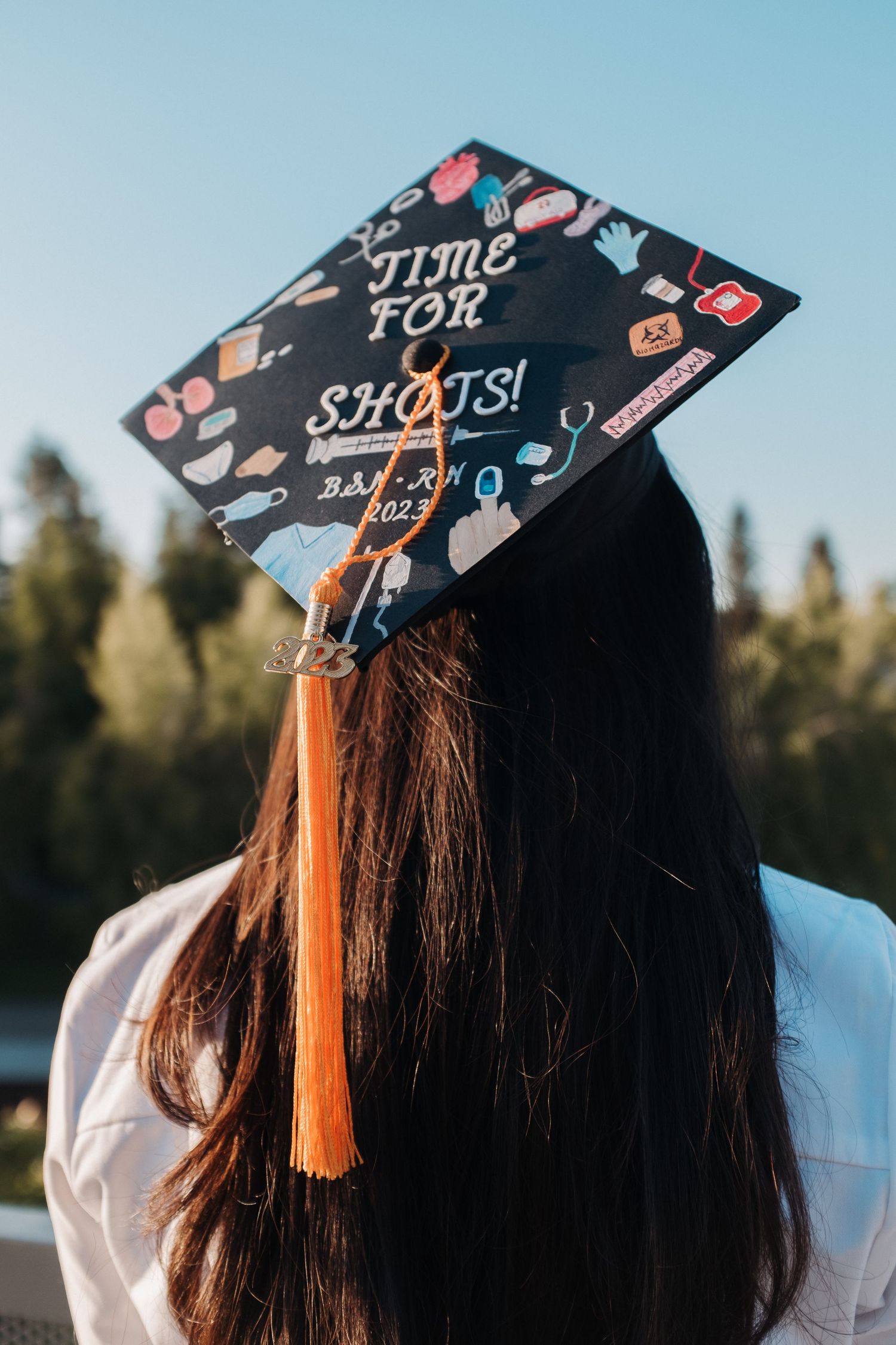 Decorated graduation cap with 'Time for Shots' design and orange tassel against blue sky.