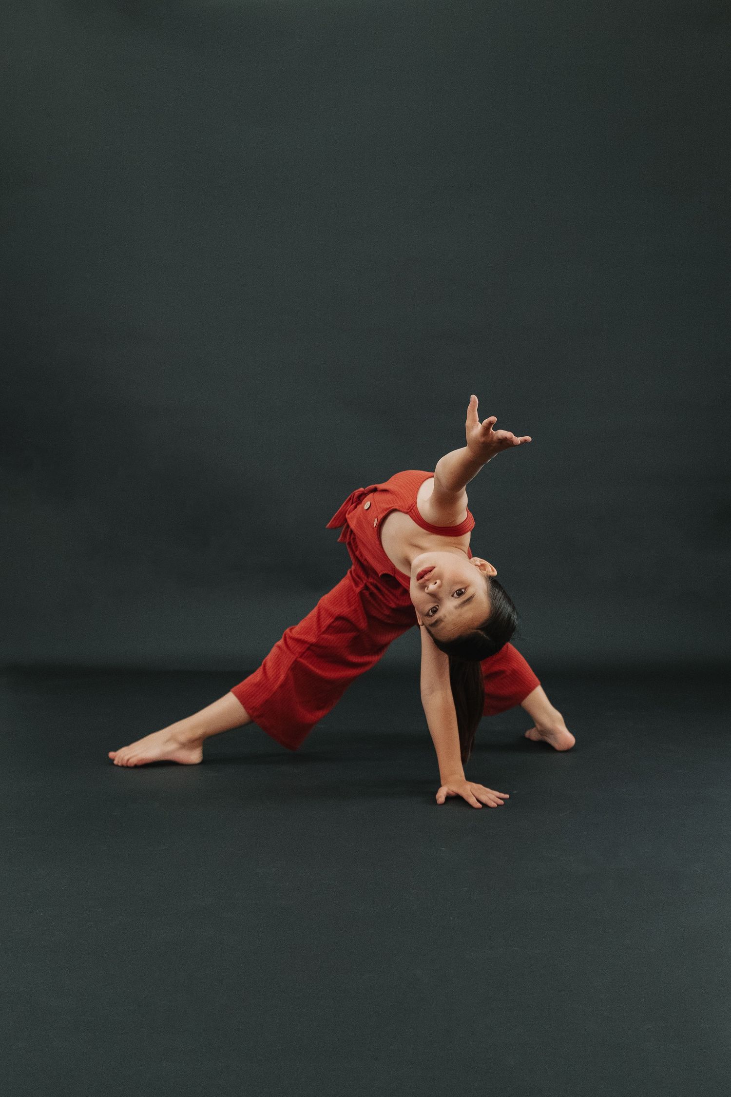 Young dancer in red outfit performs dynamic side bend on dark background.