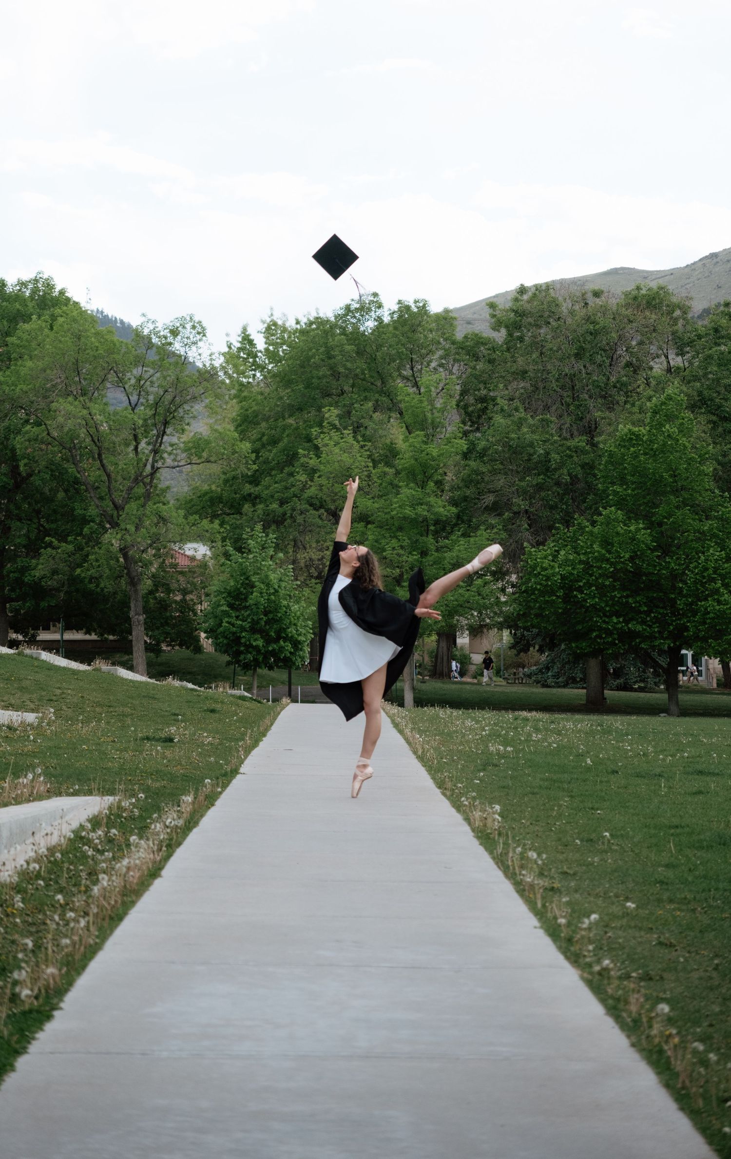 Ballet dancer in graduation gown leaps gracefully on a tree-lined path, tossing mortarboard cap into the air.