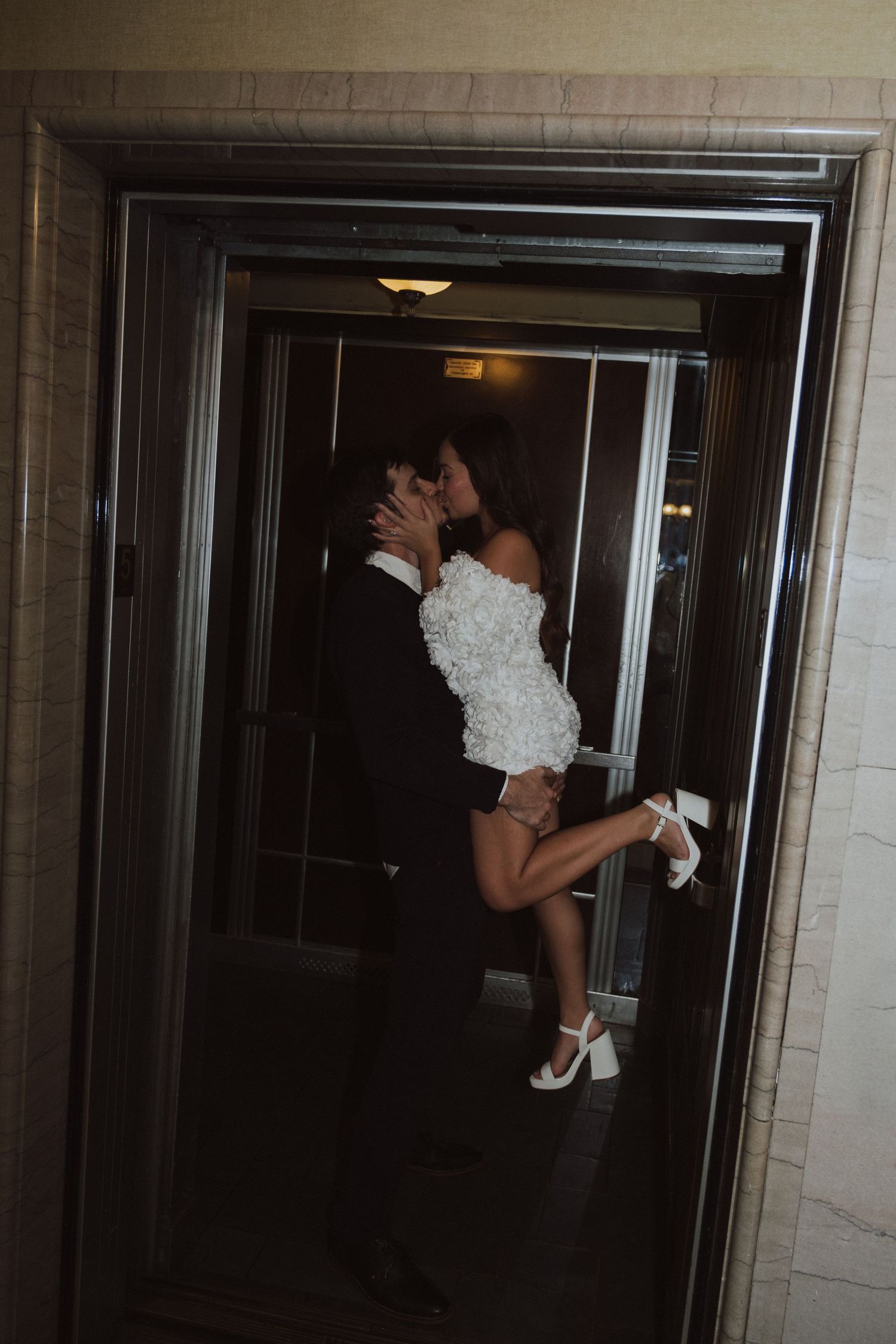 Bride and groom sharing a romantic kiss in an elevator, bride wearing a white mini dress and heels.