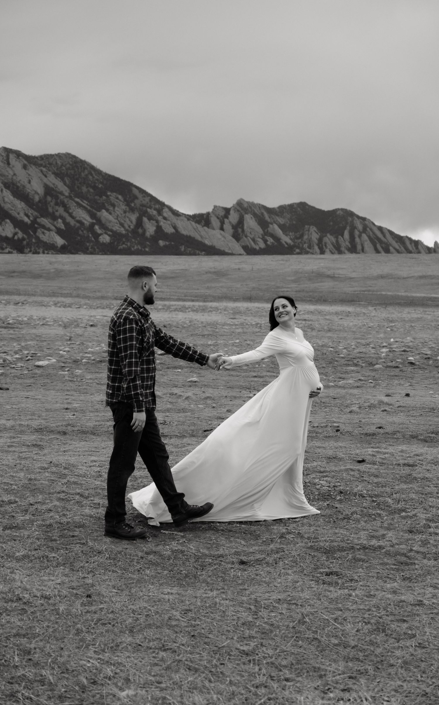 Black and white photo of a bride and groom holding hands in a field with the Flatirons mountains in the background.