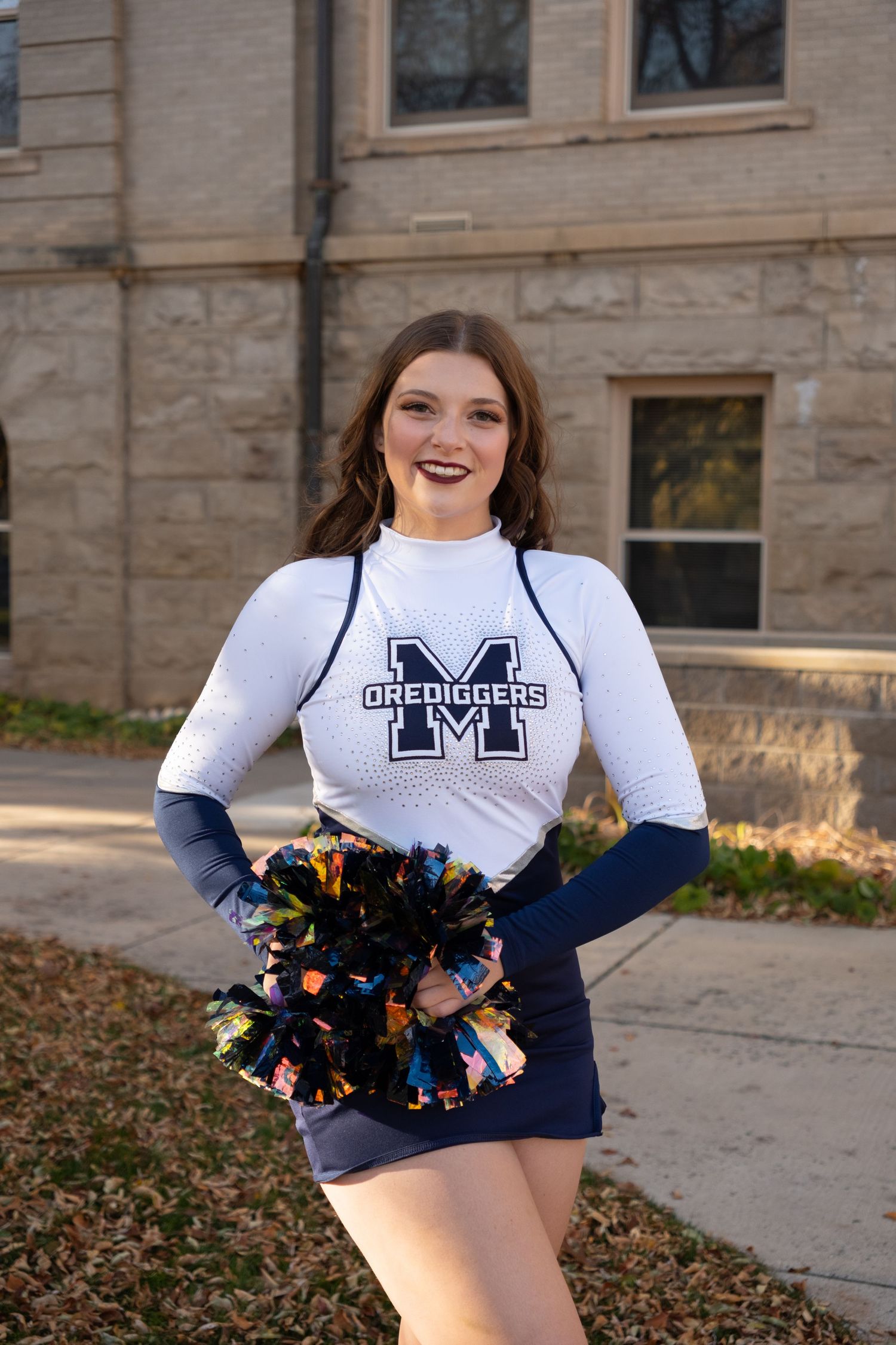 Smiling cheerleader in white M Orediggers uniform holding colorful pom-poms outdoors near a stone building.