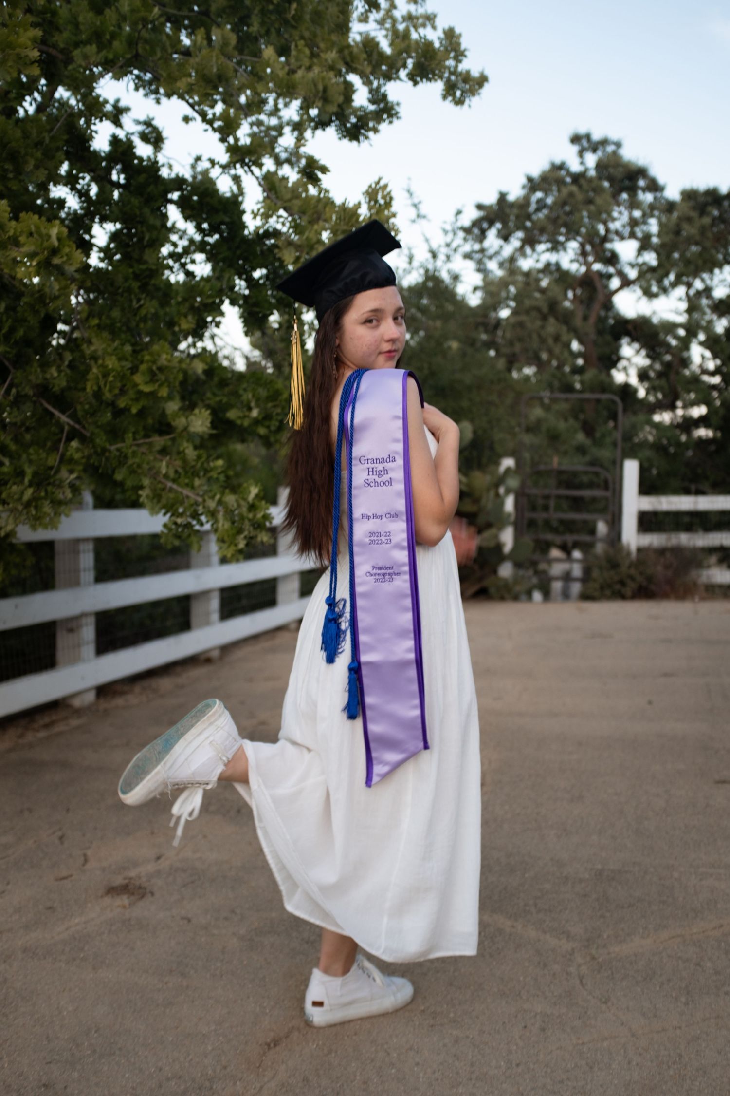 Graduate in white dress and academic regalia playfully poses outdoors.