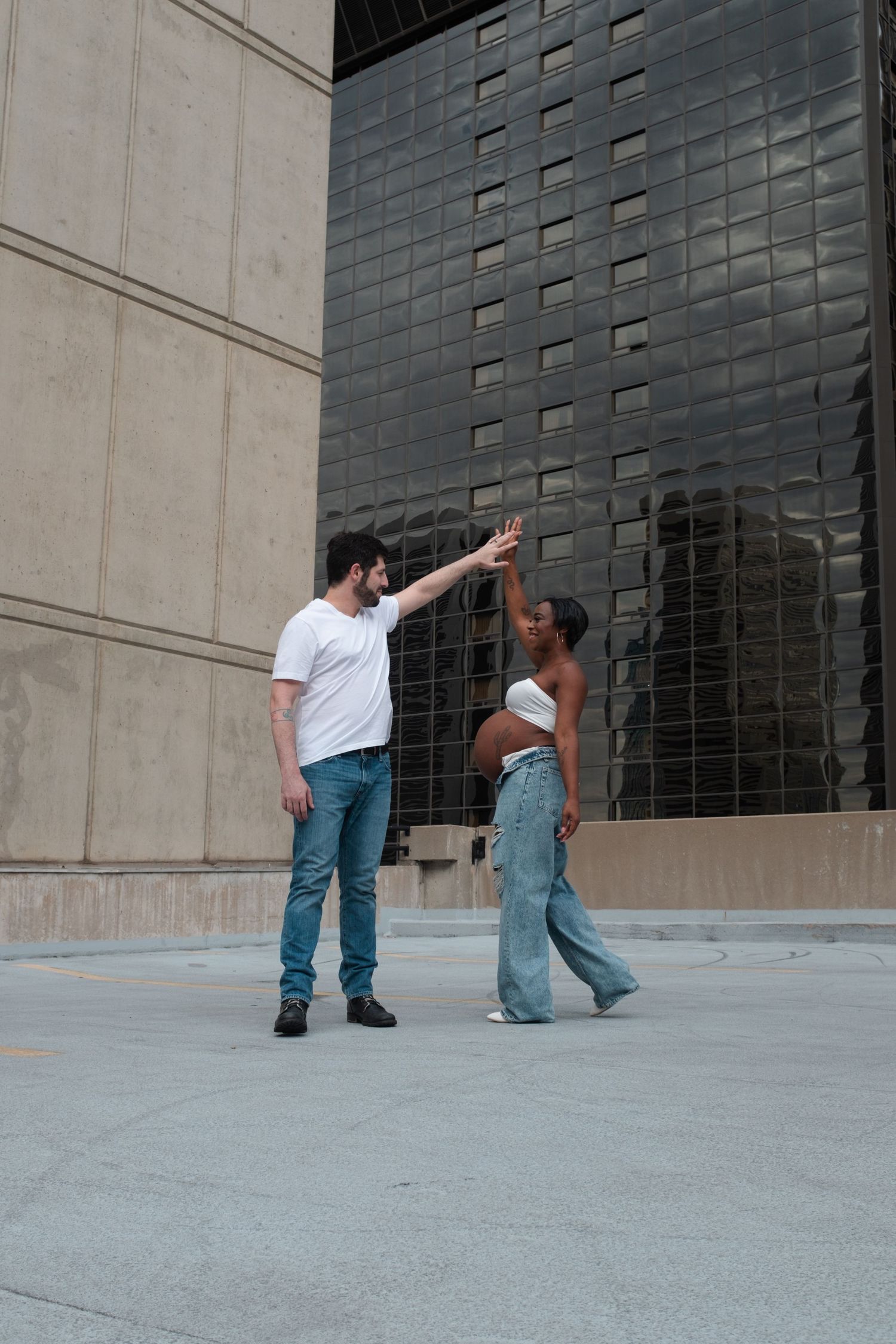 A man and pregnant woman hold hands outdoors near modern buildings in casual denim outfits.