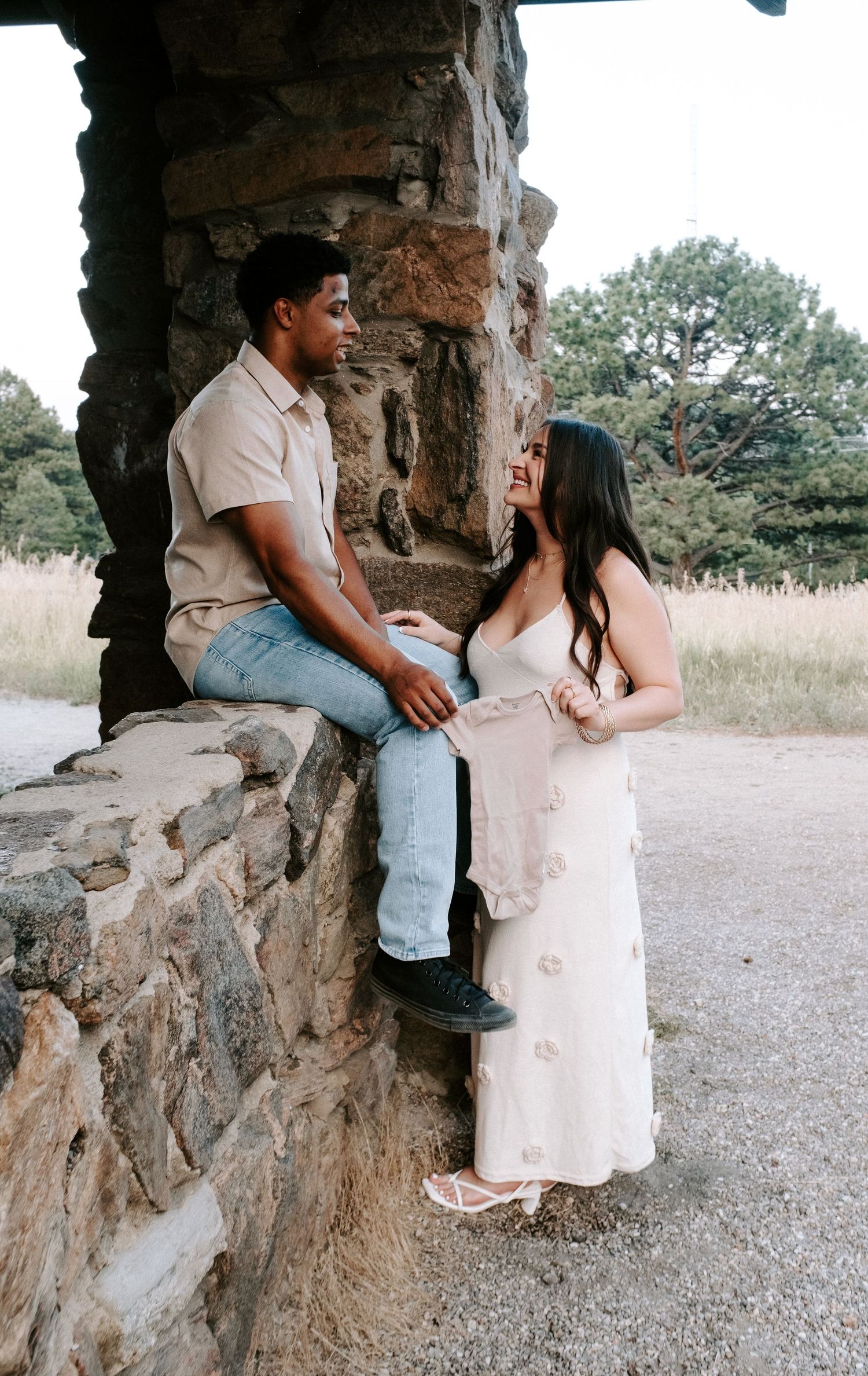 A couple shares a romantic moment by a stone structure, woman in white floral dress, man in beige shirt and jeans.