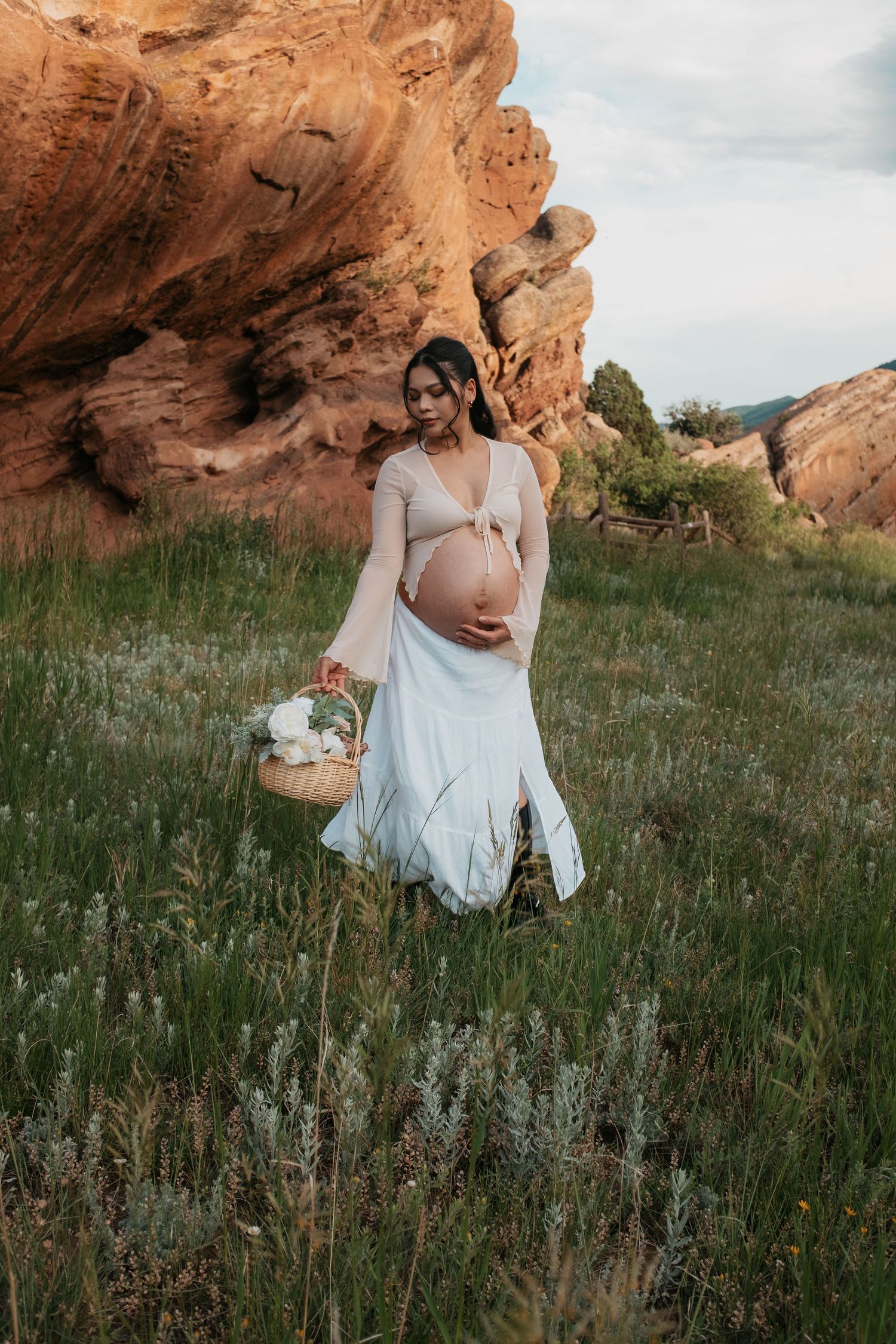 Pregnant woman in white skirt holds flower basket near red rocks in lush green meadow.