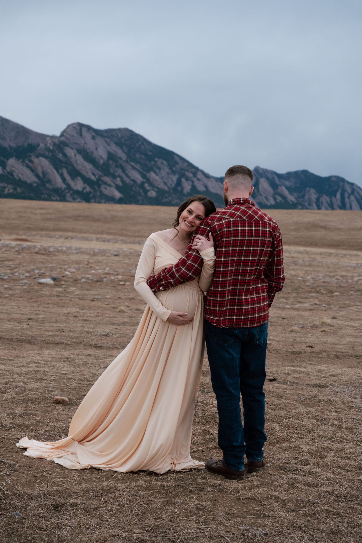 Pregnant couple embracing in a field with the Flatirons mountains backdrop, woman in flowing peach gown, man in red plaid shirt.