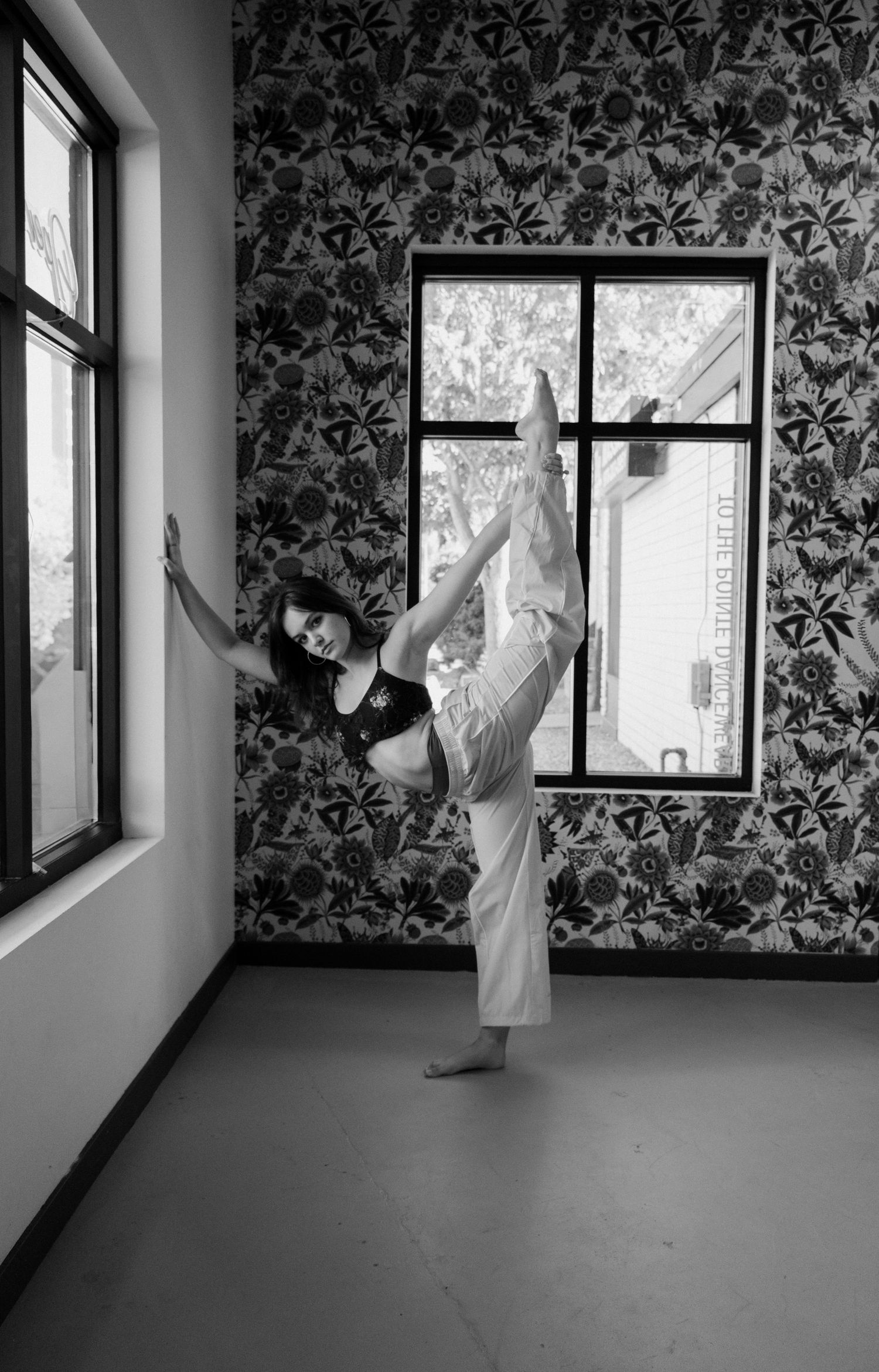 Black and white photo of a woman balancing on one leg in a dance studio with floral wallpaper.
