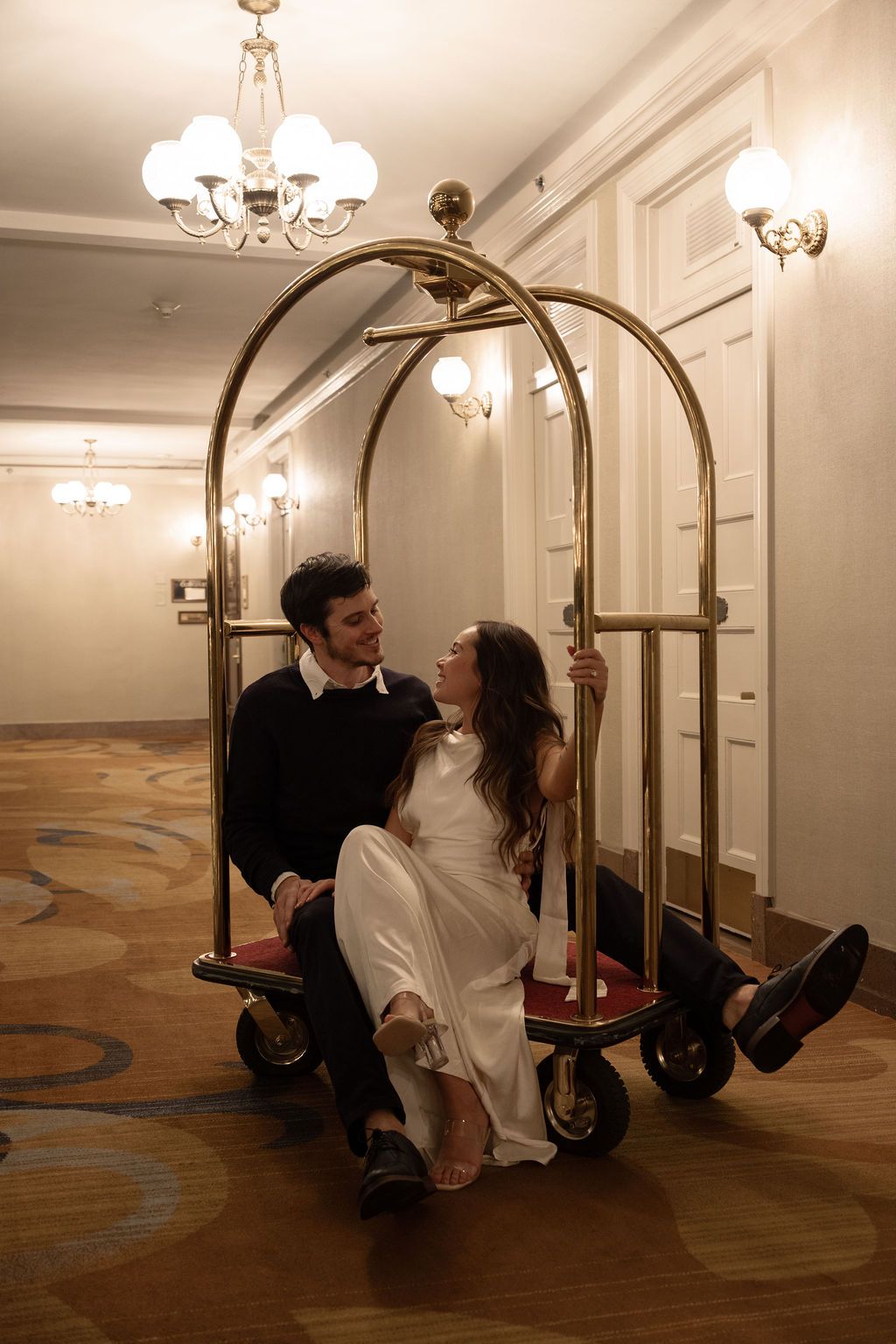 A couple sits together in a gold hotel luggage cart in an elegant hotel corridor with chandeliers.