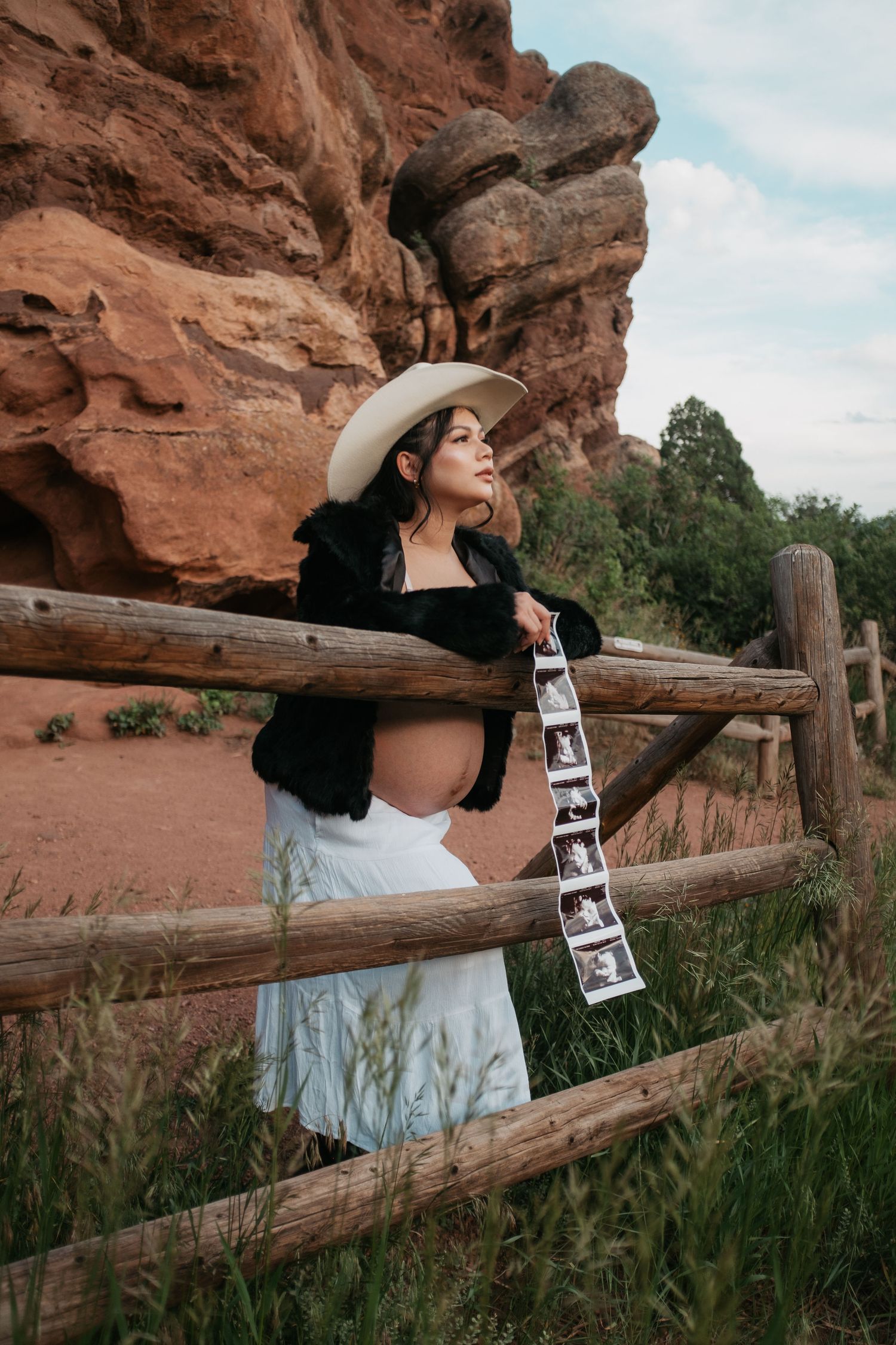 Pregnant woman in cowboy hat and black jacket holds ultrasound photos by rustic fence at Red Rocks, Colorado.