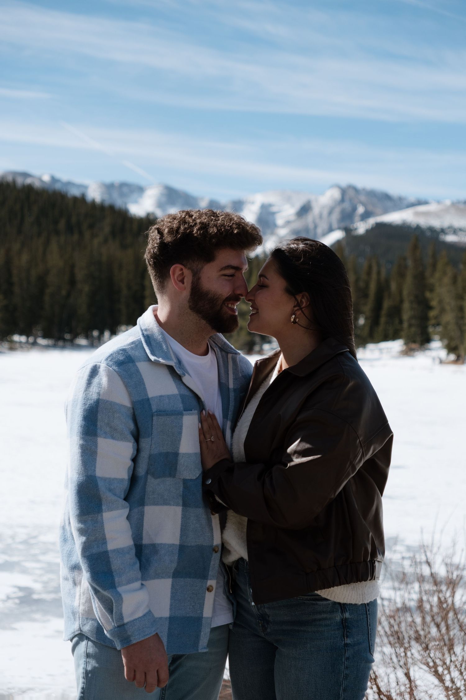 Couple sharing a romantic moment in a snowy mountain landscape with pine trees and blue skies.