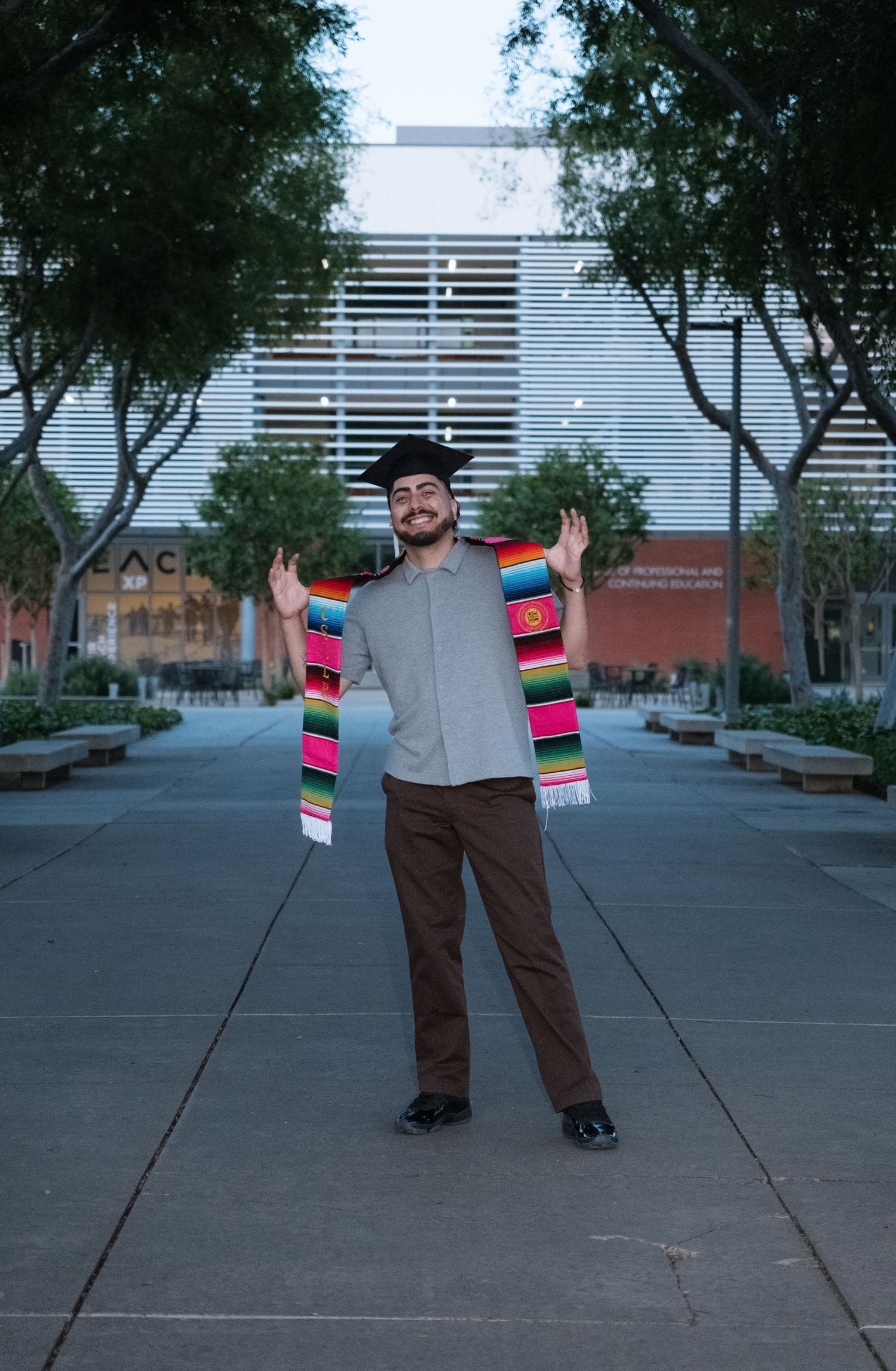 Graduate in cap and colorful serape scarf poses with arms raised outdoors near a modern building.