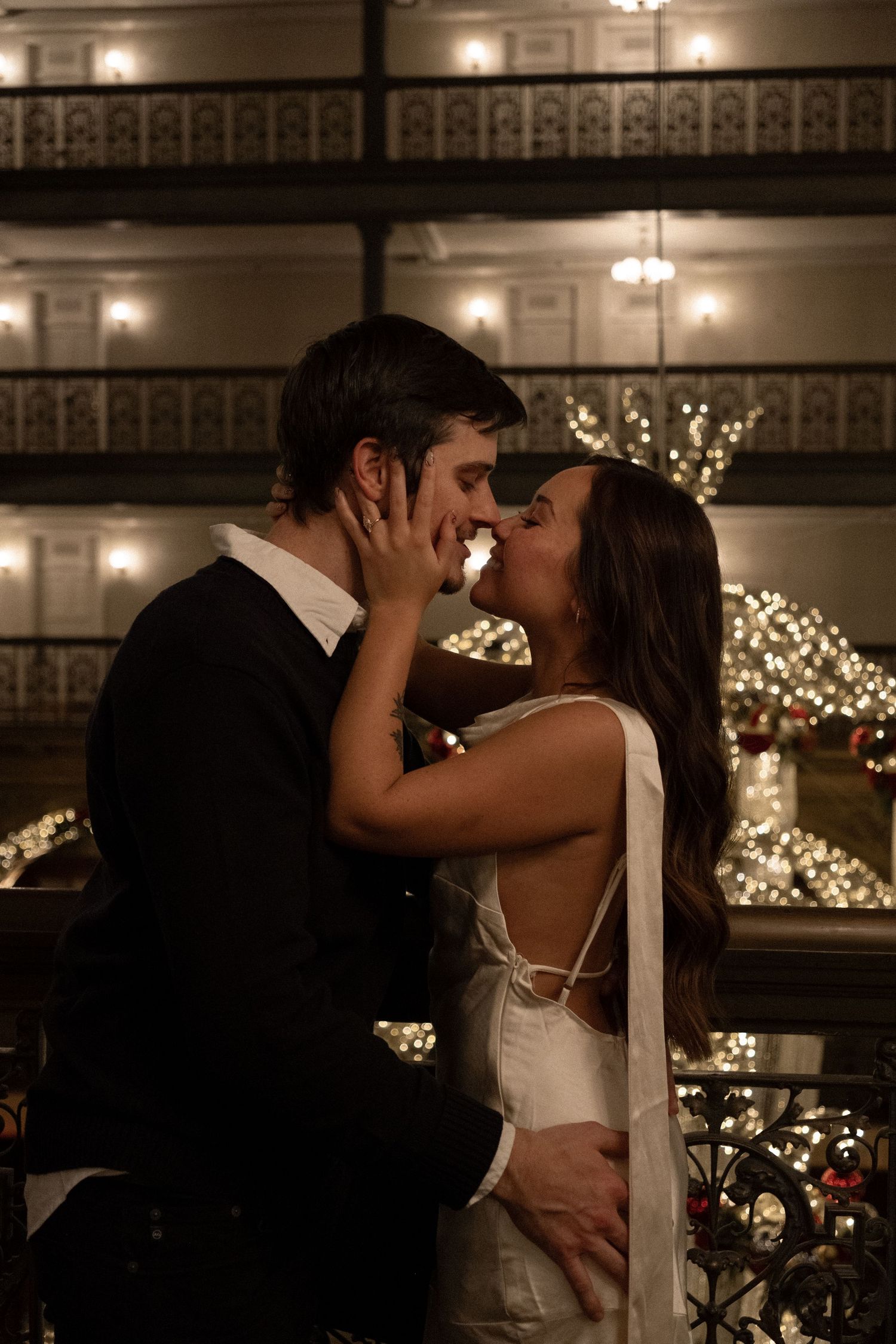 Couple sharing a romantic kiss in wedding attire surrounded by warm twinkling lights at night.