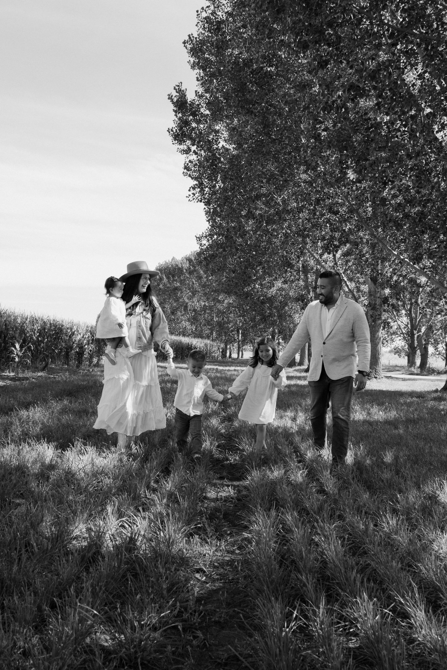 Black and white photo of a family walking together through a tree-lined field, parents and children dressed in white.